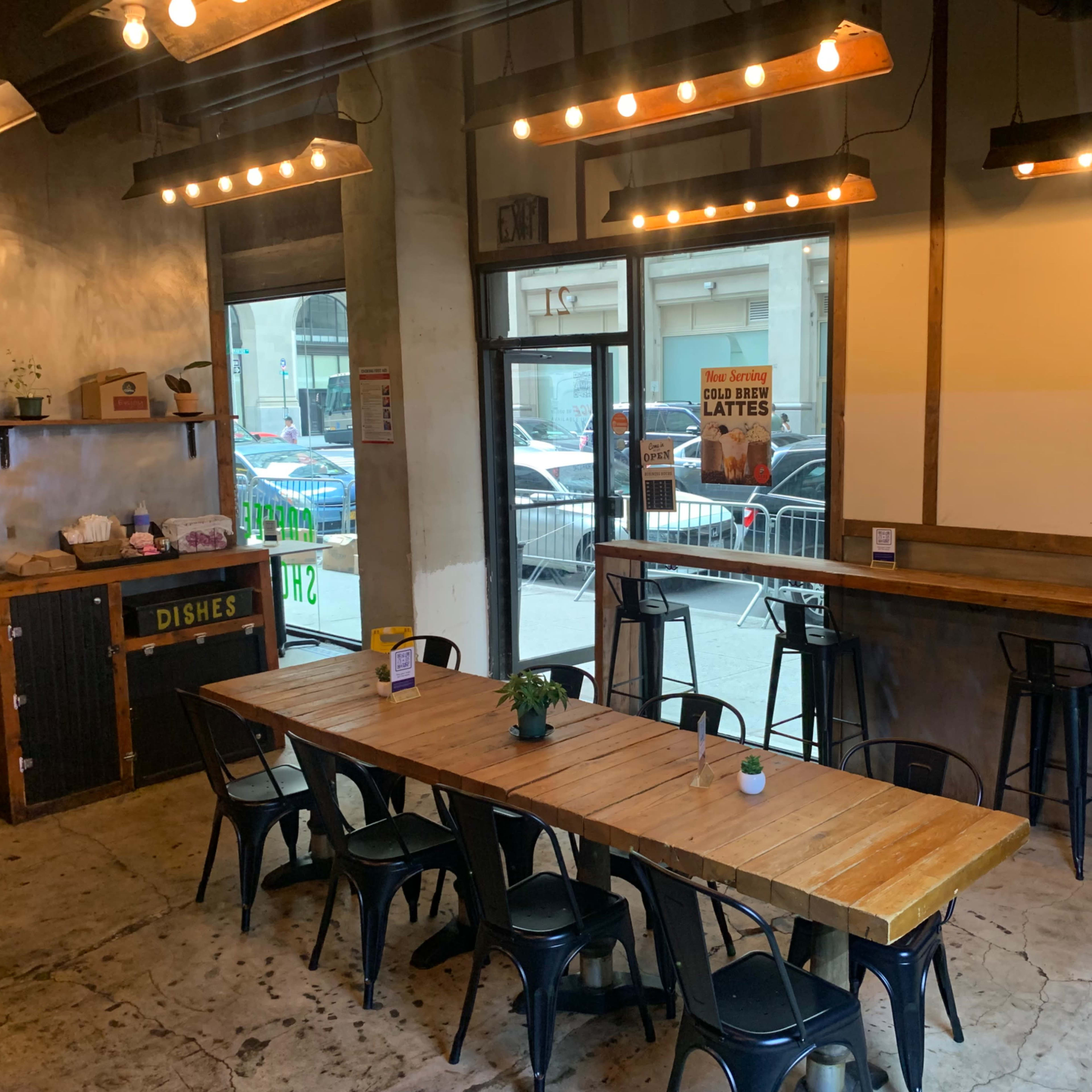 A spacious café interior featuring a long wooden dining table surrounded by black metal chairs, with large windows allowing natural light and a view of the street outside.