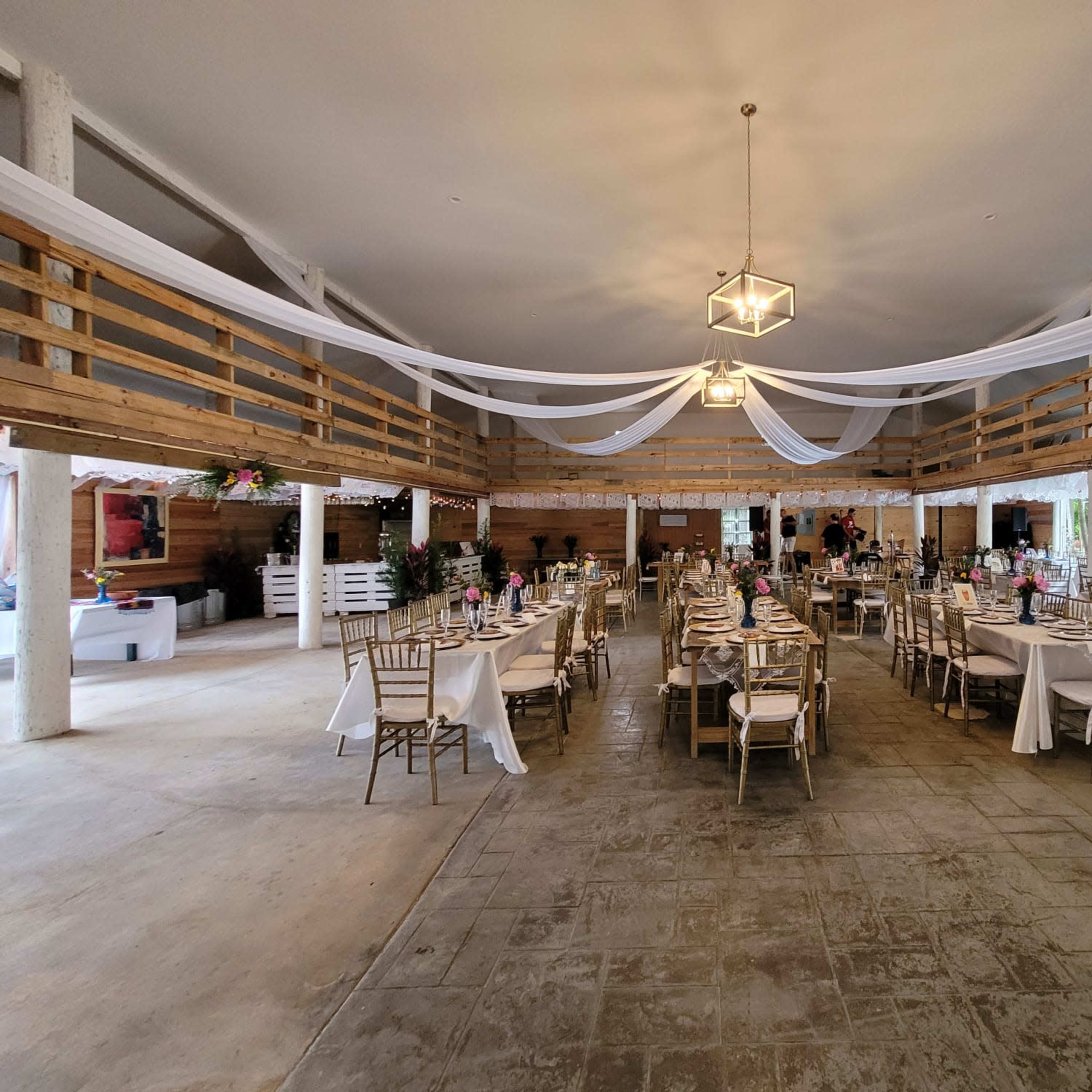 A spacious indoor event venue with neatly arranged tables covered in white linens and decorations, featuring a rustic wooden decor and a high ceiling with draped fabric.