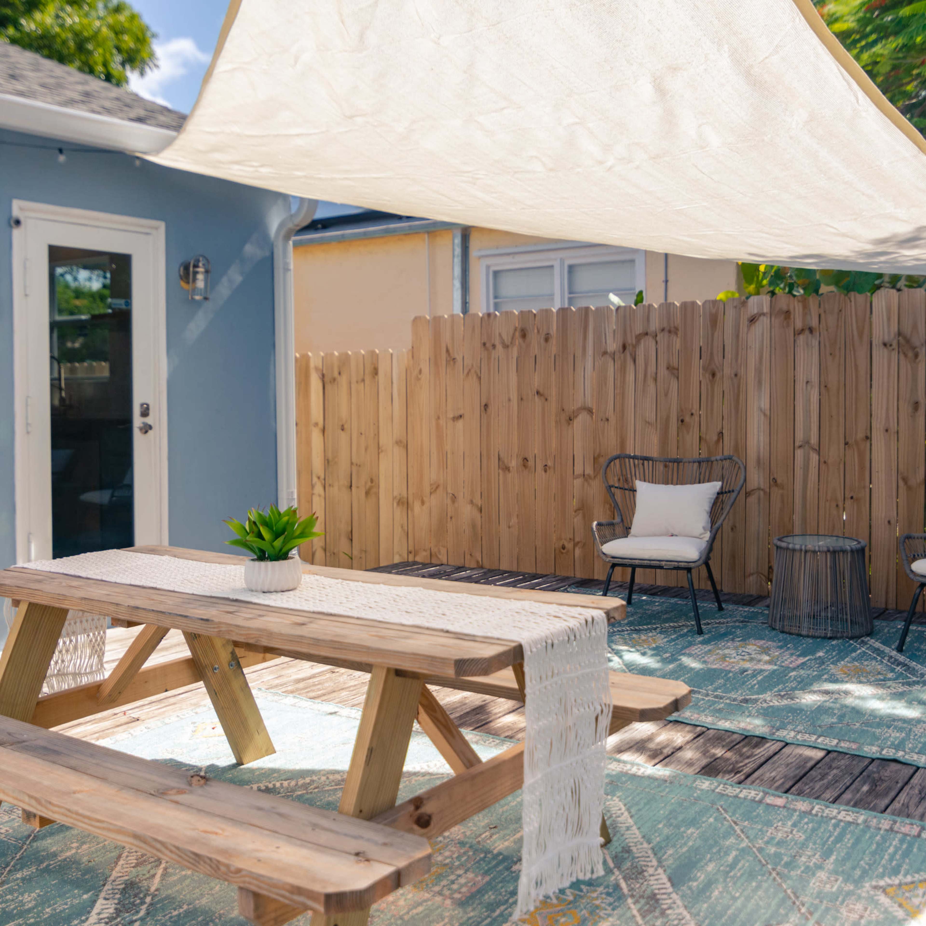 A wooden picnic table under a shaded area with two chairs and a small table on a wooden deck surrounded by a fenced yard.