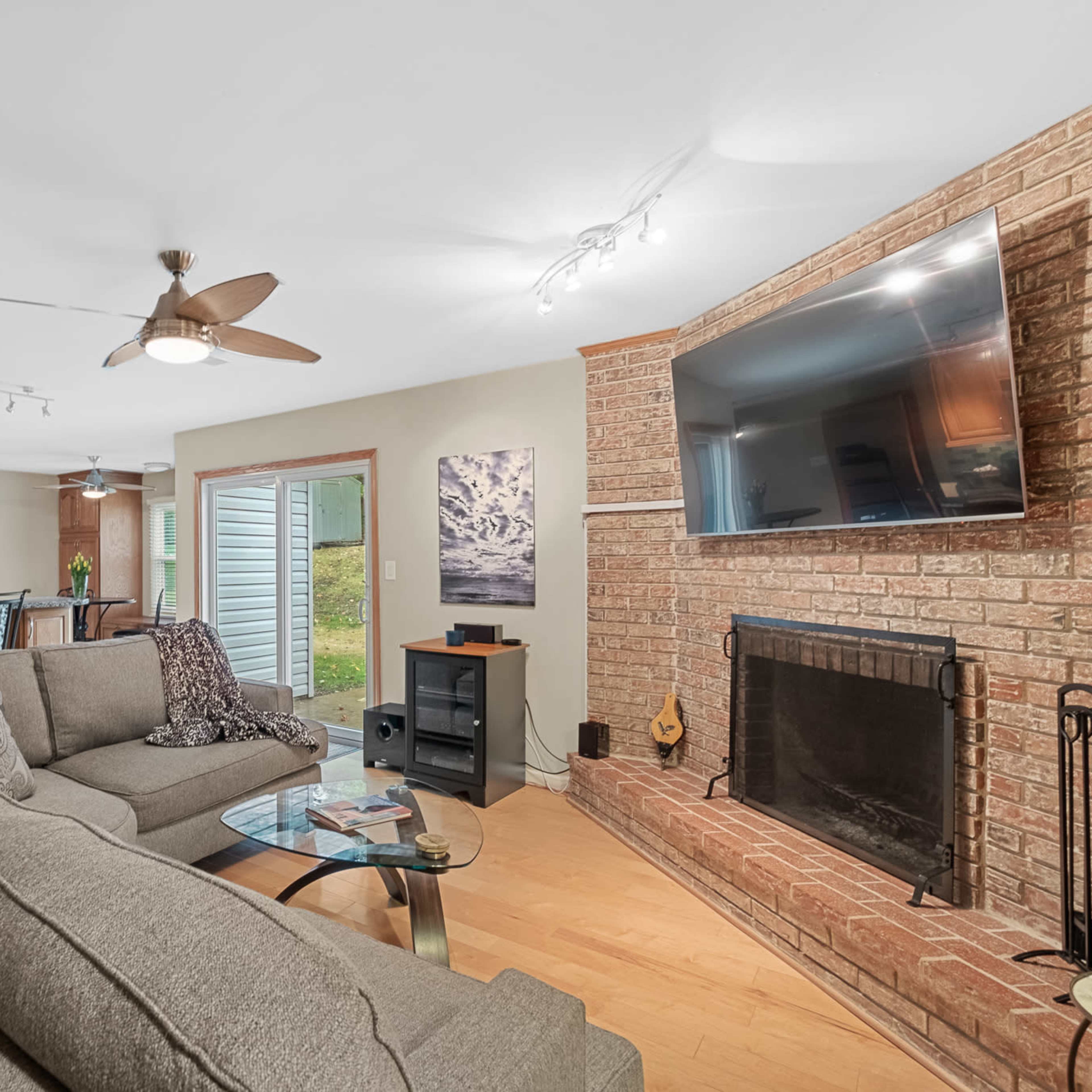 A living room with a grey sectional sofa, a brick fireplace, and a television mounted on the wall, adjacent to an open kitchen area with wooden cabinetry.