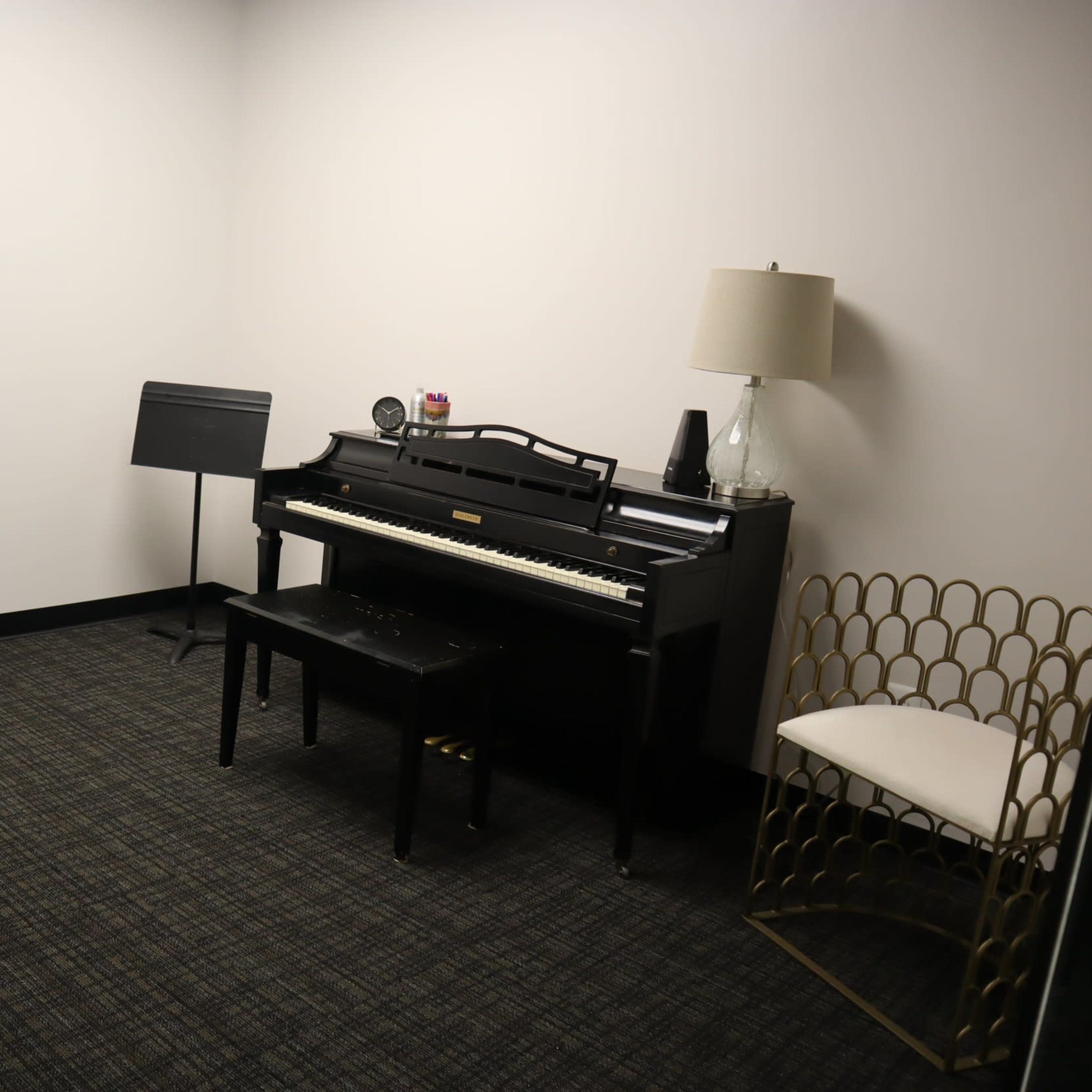 A small room with a black piano, a music stand, two black chairs, and a gold decorative chair beside a lamp.