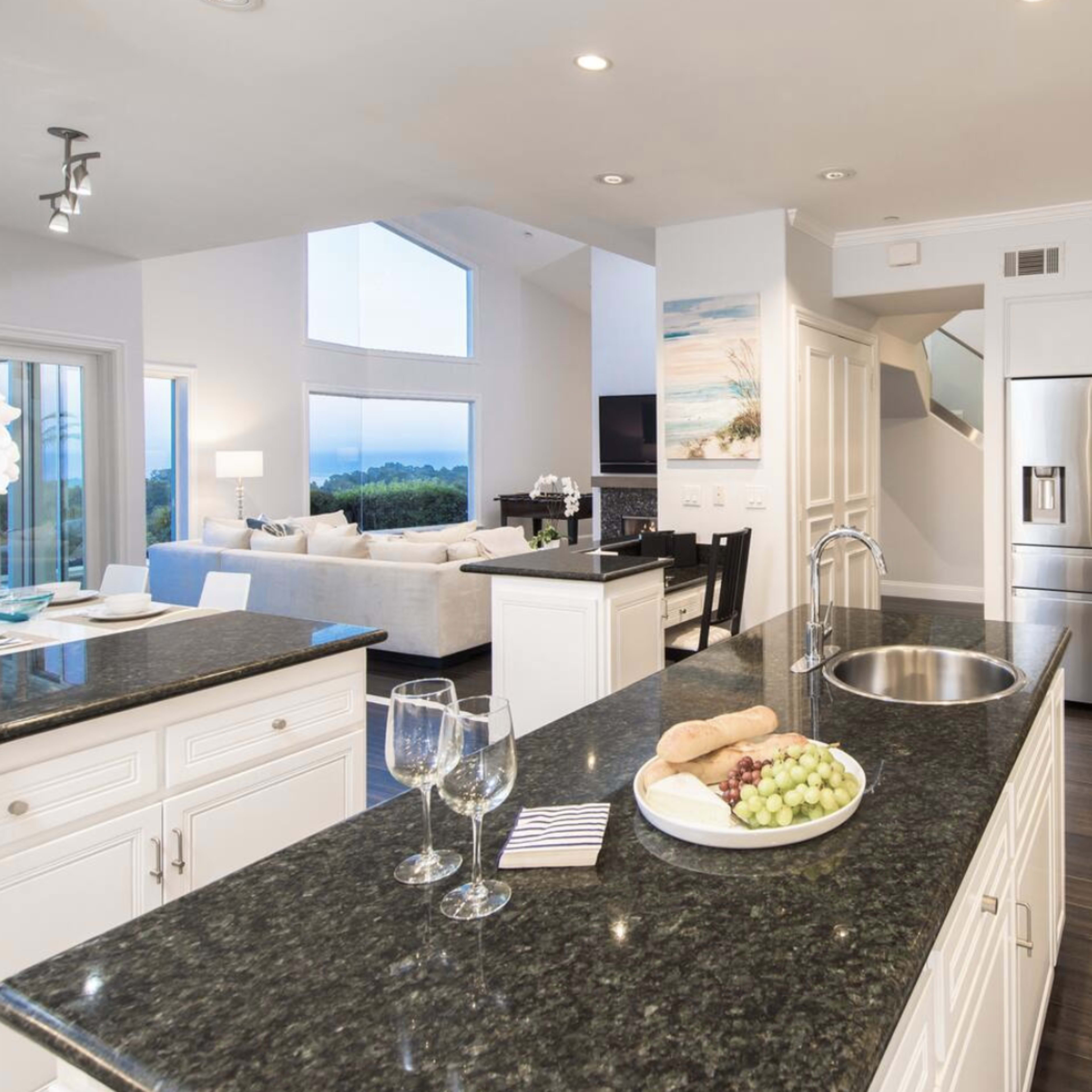 A modern kitchen featuring a dark granite countertop with a fruit platter, stainless steel appliances, and an open layout leading to a living area.