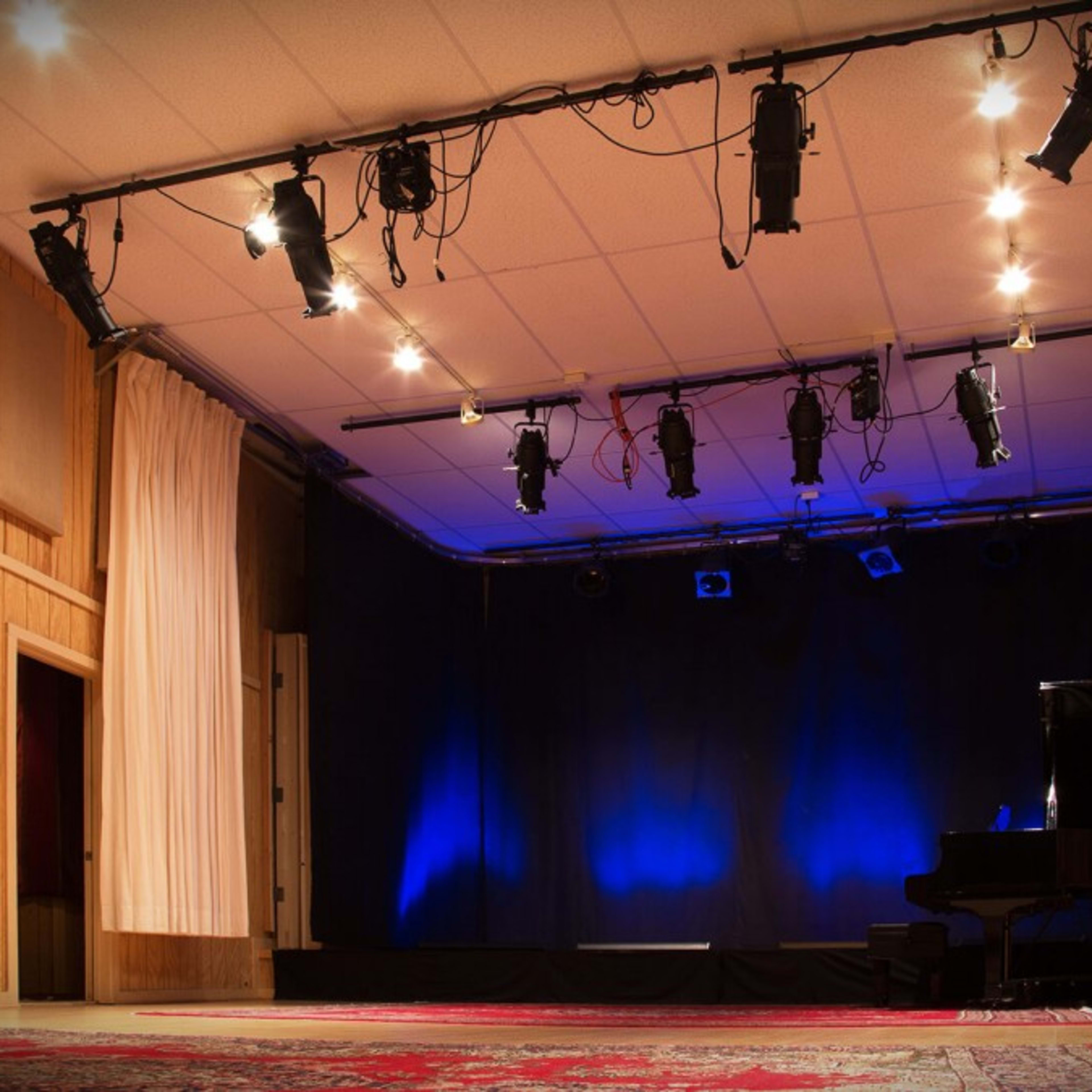 A sparsely decorated stage with a grand piano and blue lighting, set in a room with wooden paneling and soundproofing panels.