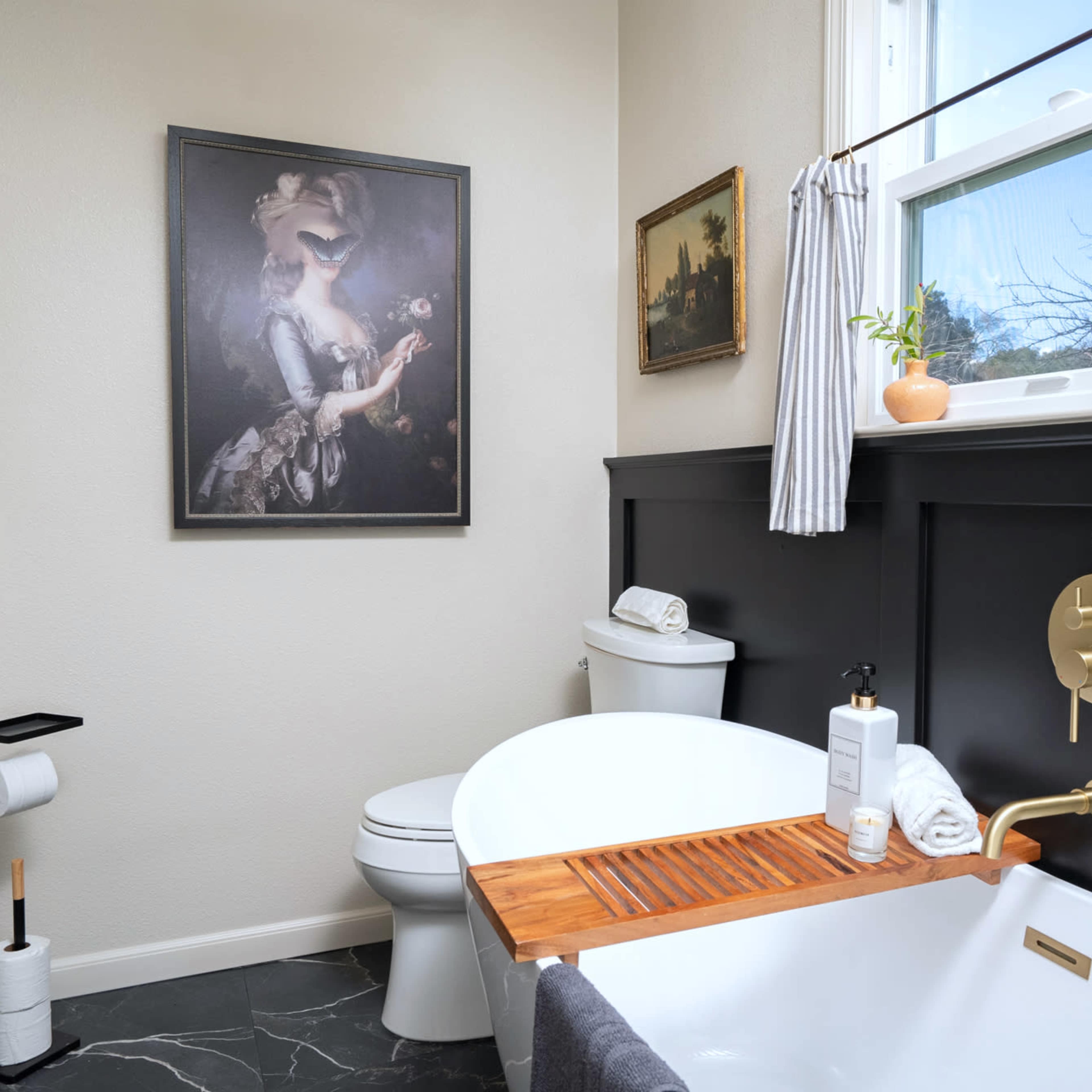 A bathroom featuring a freestanding bathtub, a toilet, and framed artwork on the walls, with dark wainscoting and a large window.