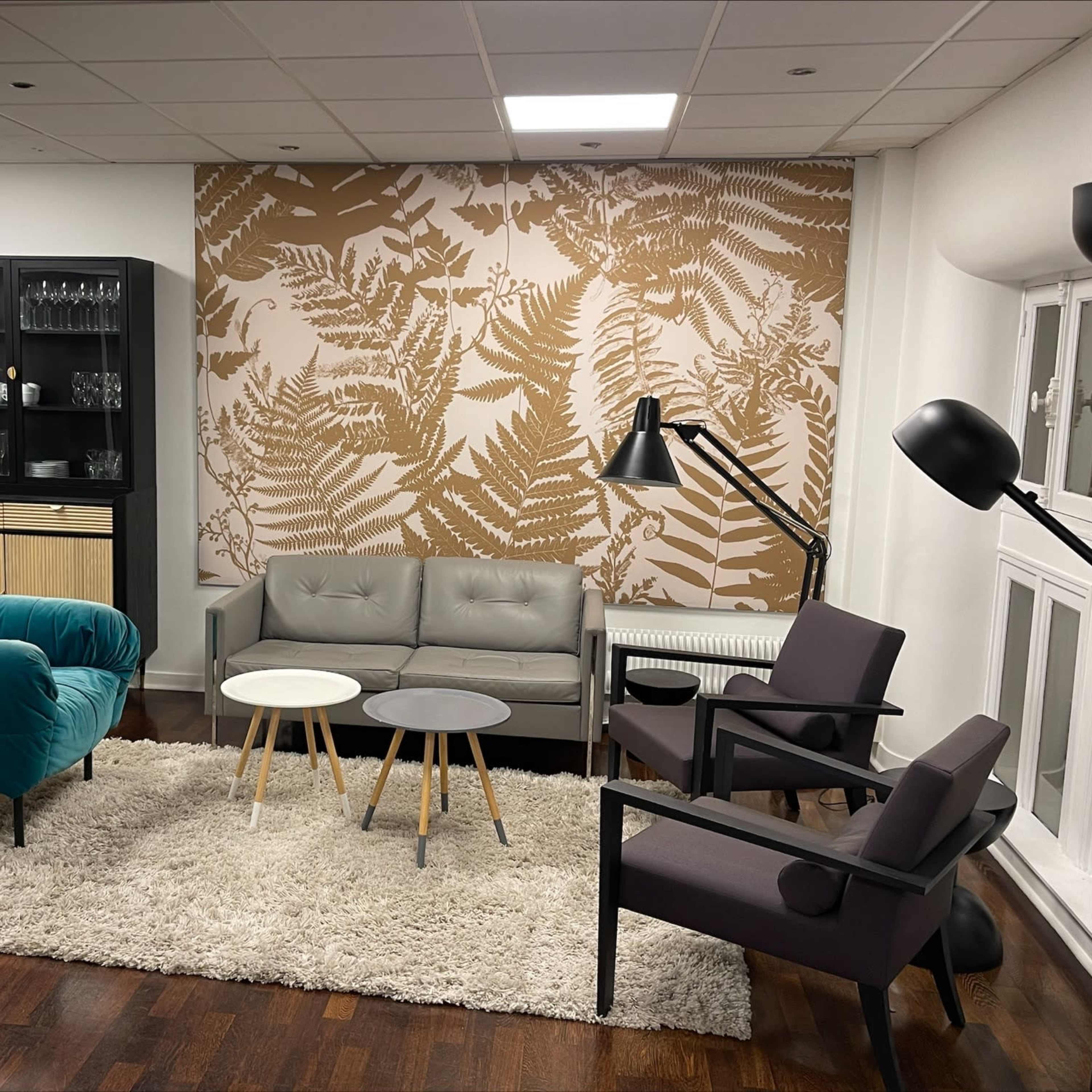 A modern lounge area with a teal sofa, gray armchairs, a coffee table, and a patterned wall featuring fern designs.