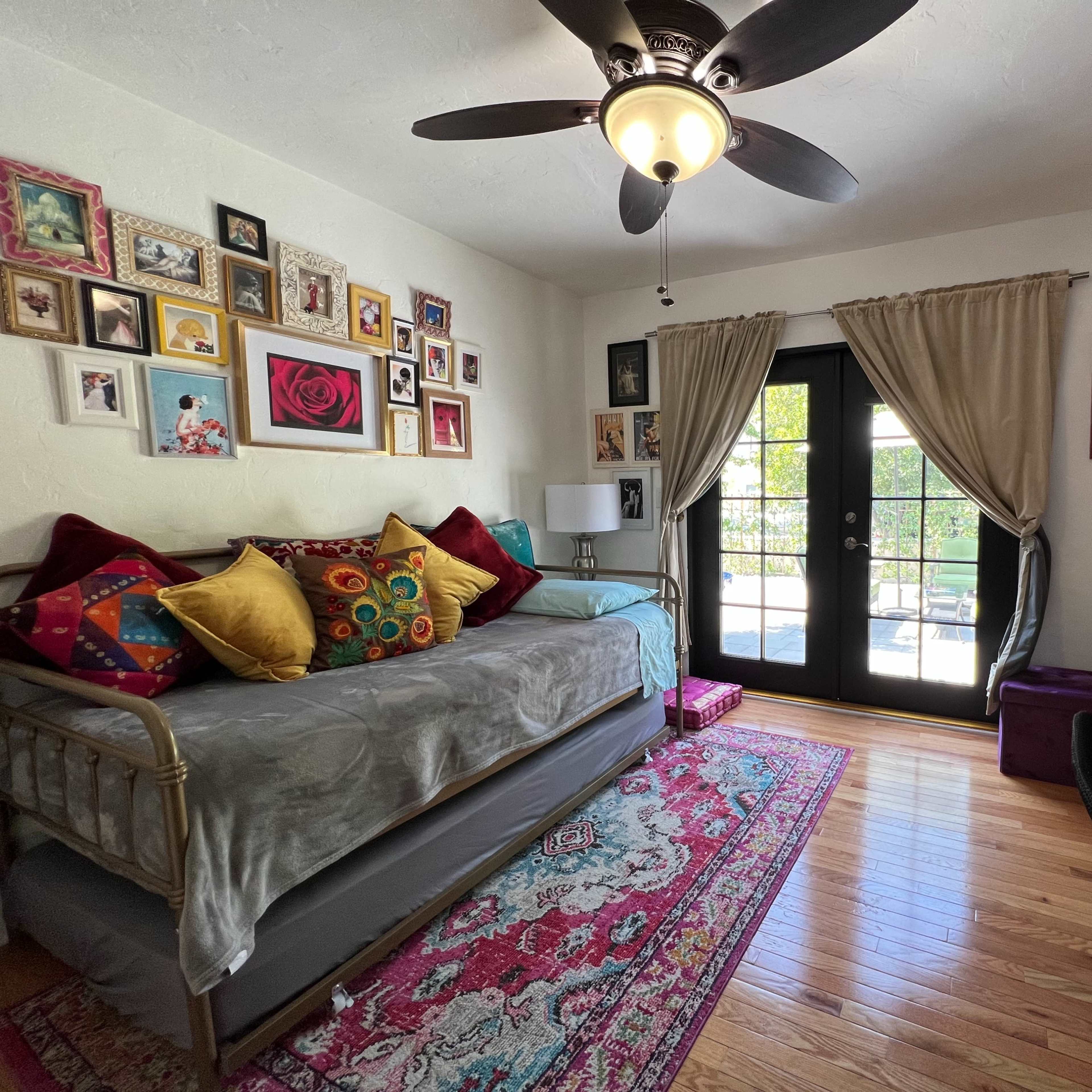 The room features a daybed adorned with colorful pillows, a desk against the wall, and large windows with double doors leading to an outdoor space.
