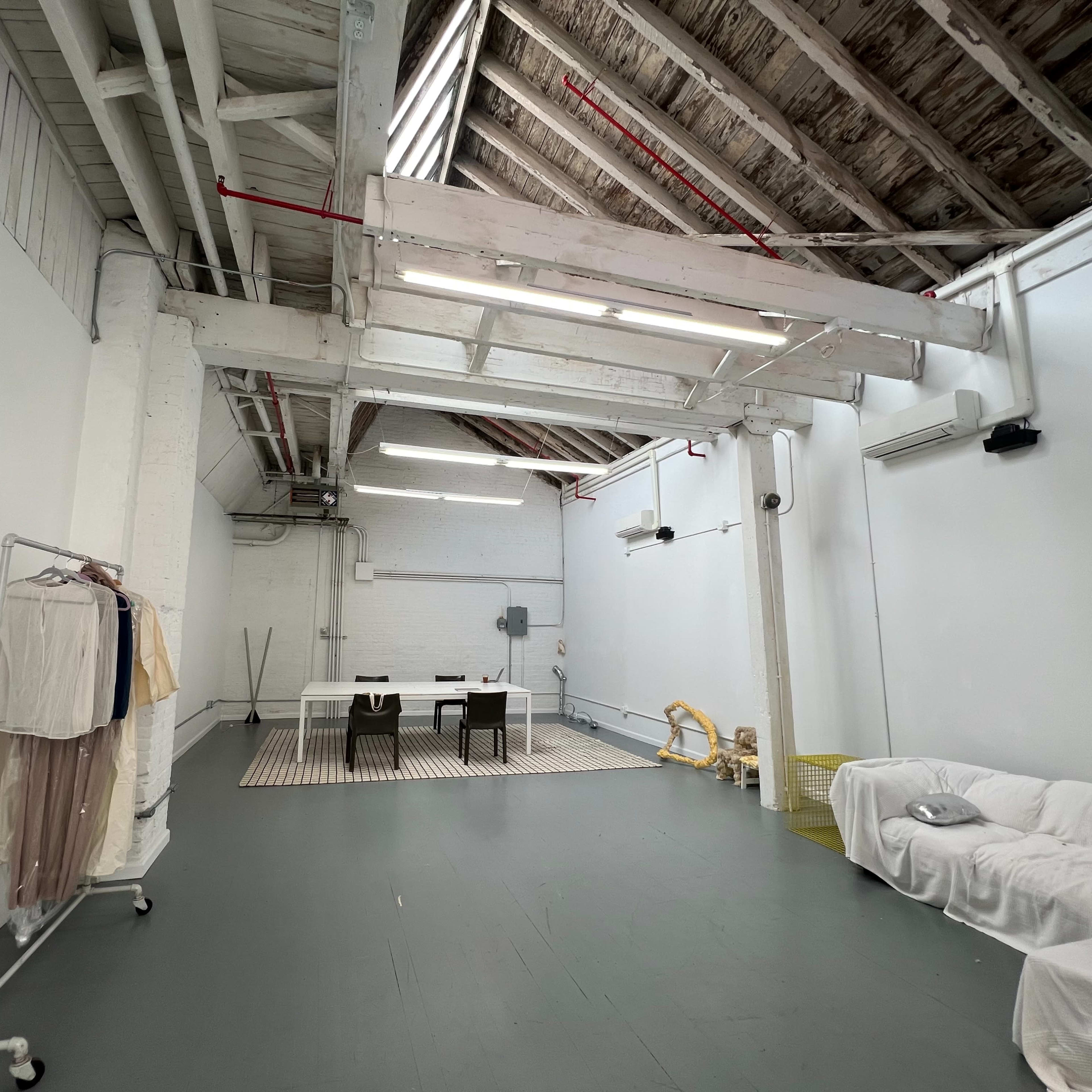 A spacious, minimalist interior with exposed beams, a large table, chairs, and a clothing rack against the white walls.