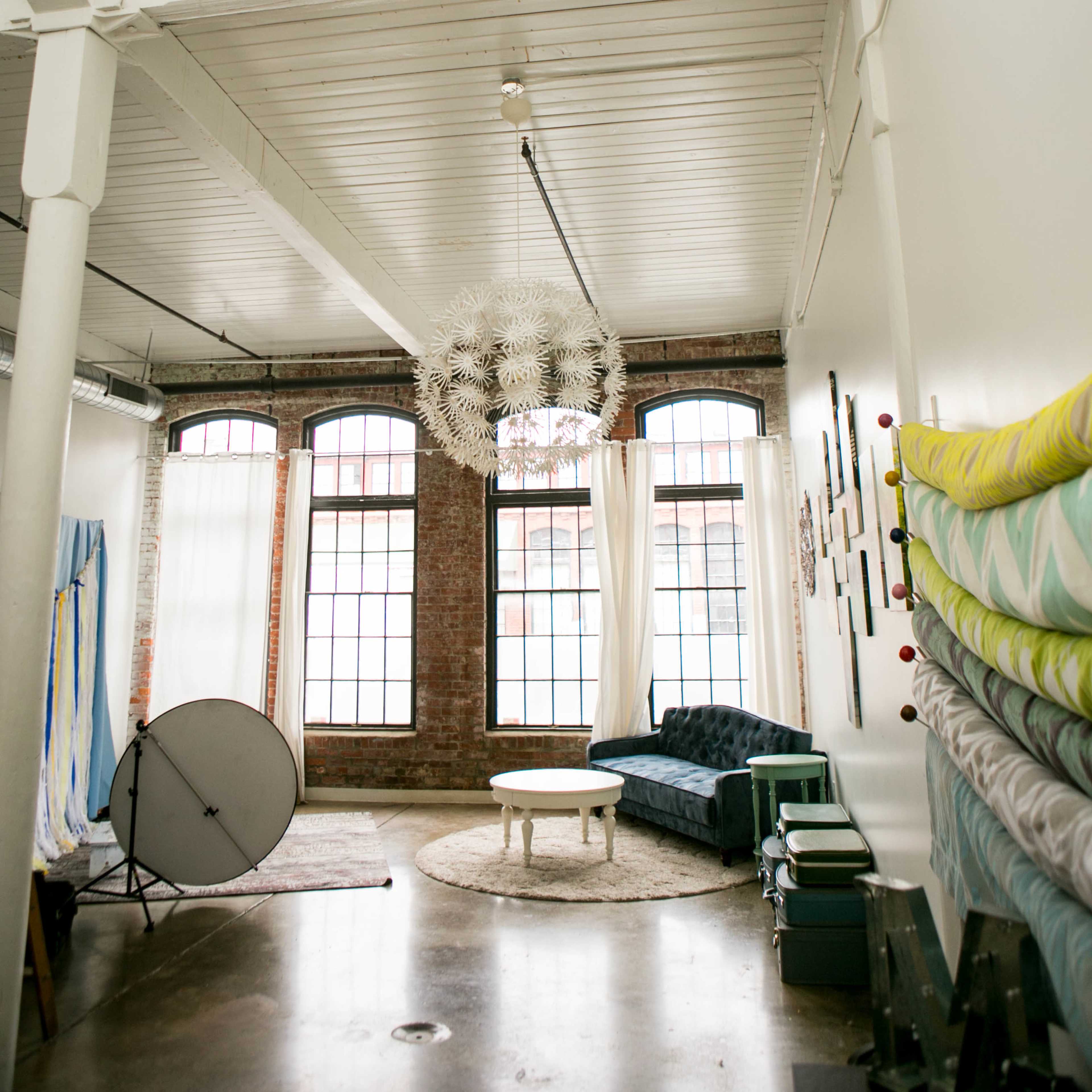 A spacious, well-lit interior with a high ceiling, large windows, and a central circular rug beneath a coffee table, surrounded by a sectional sofa and decorative wall hangings.