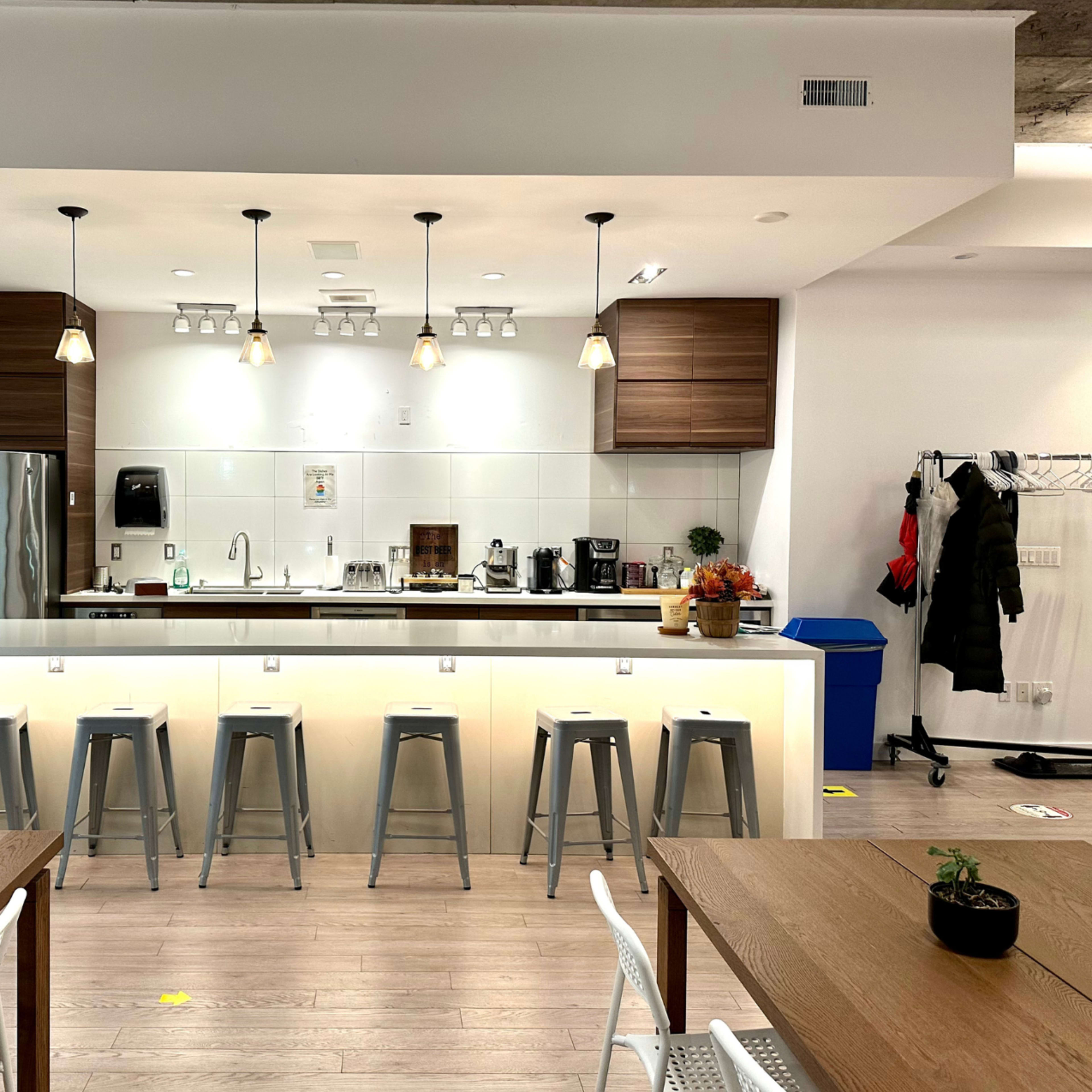 A modern kitchen area with a bar counter, stools, and various appliances, including a refrigerator and coffee machines.