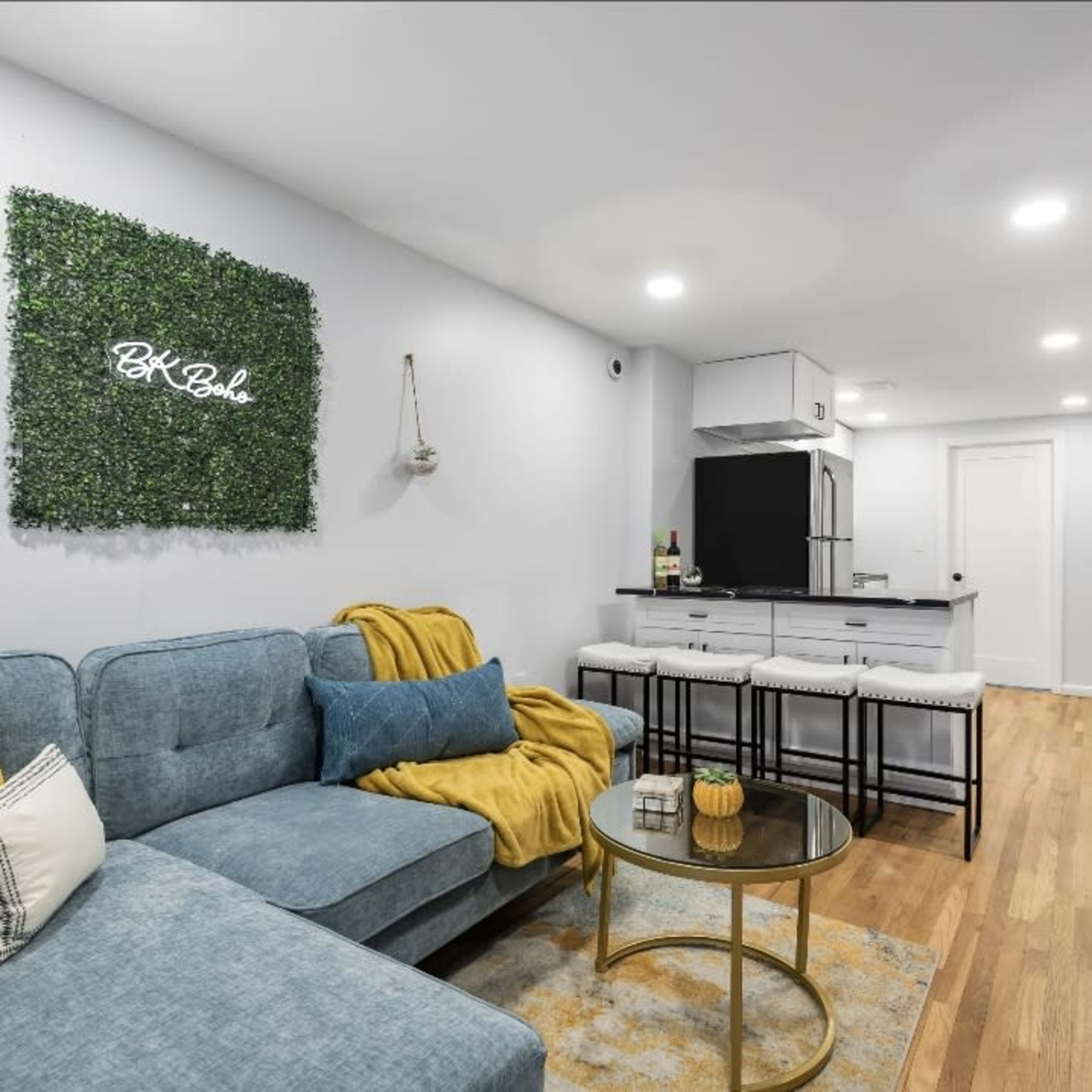 A modern living space with a light-colored wall, a blue sofa adorned with yellow and gray pillows, a round coffee table, and a dining area with barstools next to an open kitchen.