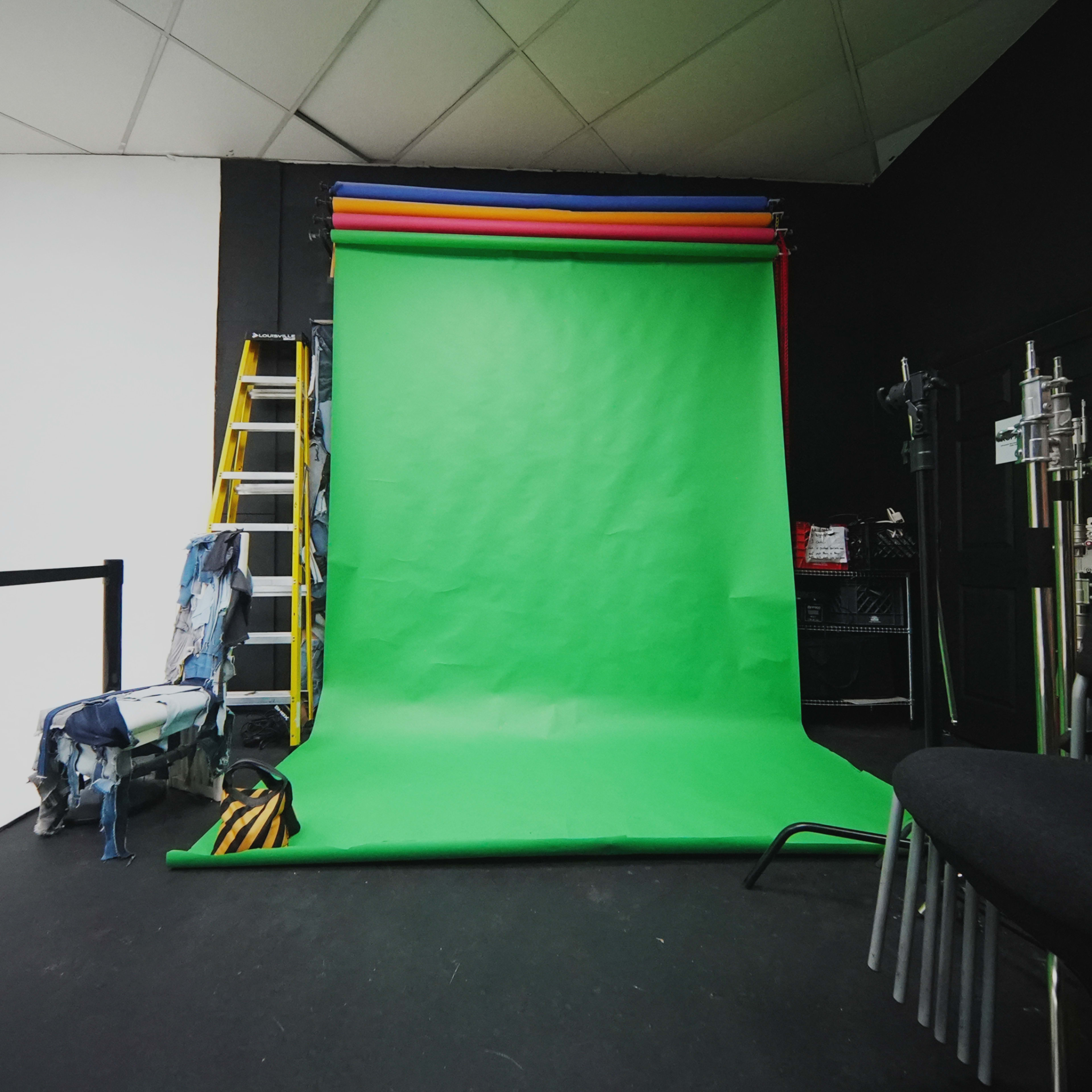 A photo studio featuring a green backdrop, a ladder, and various equipment along with a stack of chairs.