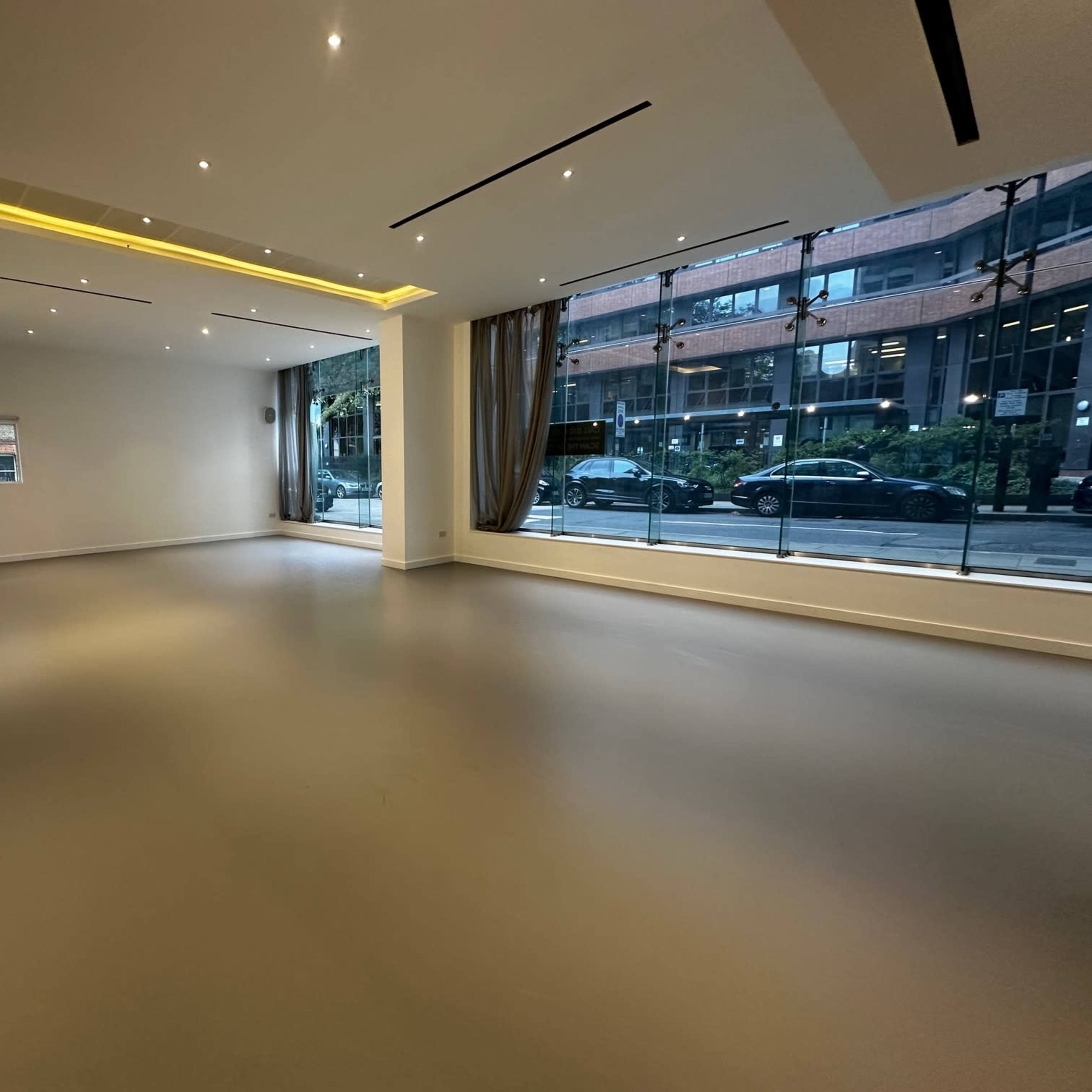 A large, empty room with polished flooring and ample natural light from large windows facing the street.