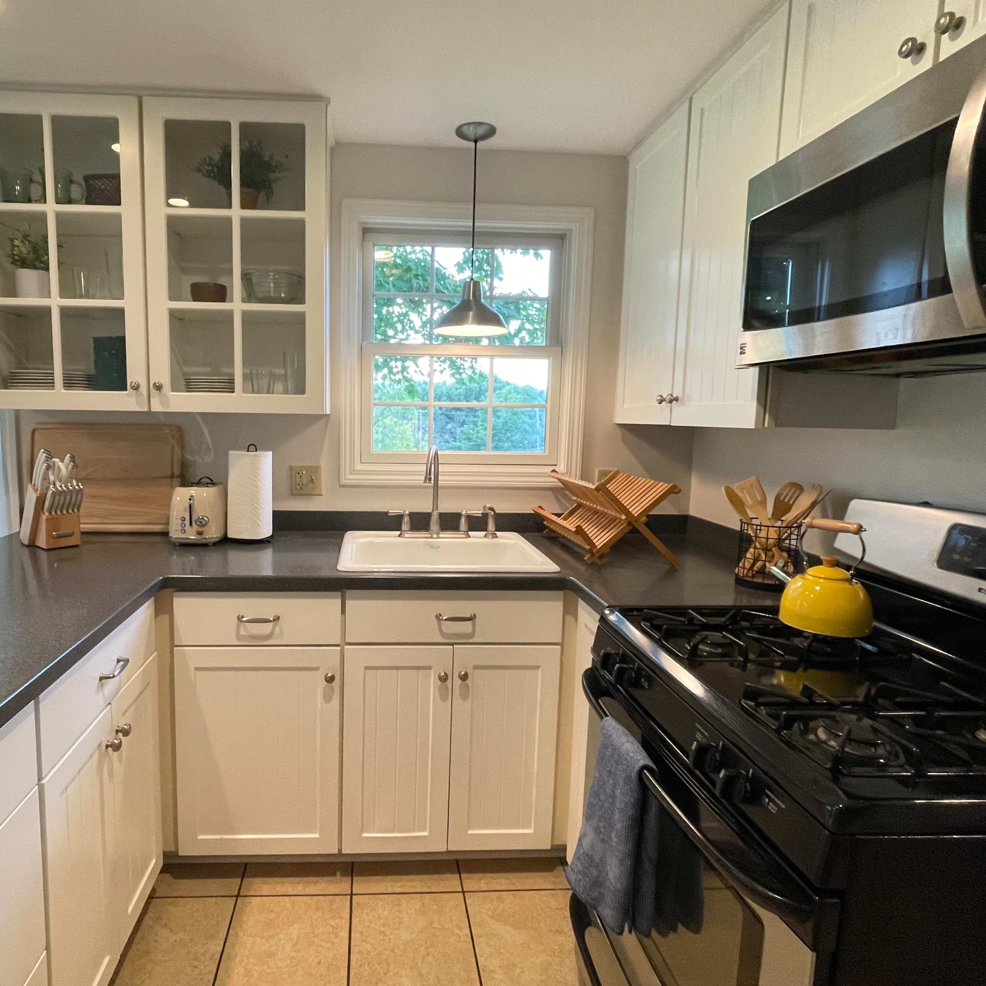 A modern kitchen features white cabinetry, a stainless steel microwave, and a gas stove, with a sink beneath a window overlooking a green outdoor view.