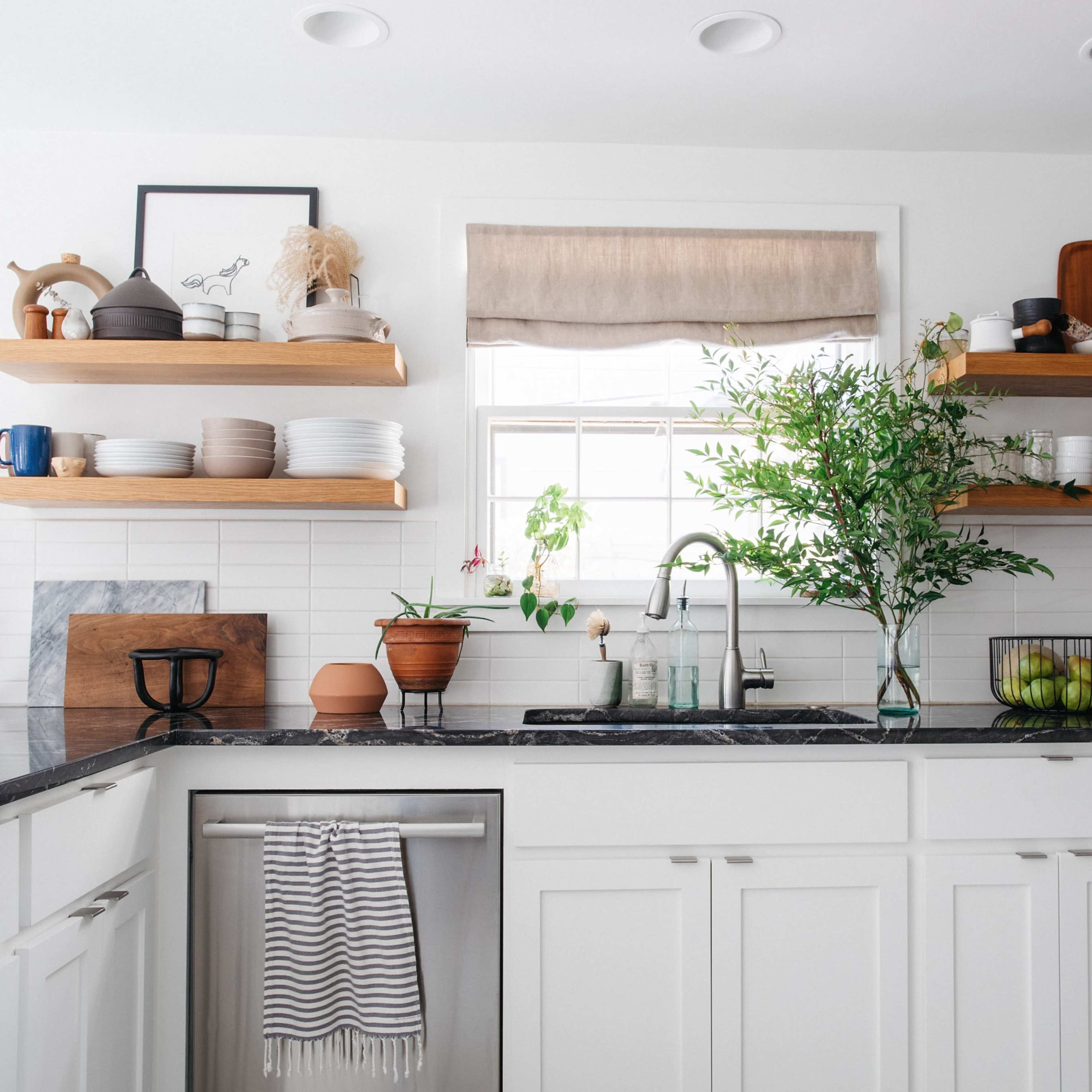 A modern kitchen with white cabinetry, open wooden shelves filled with dishes, and a sink under a window with potted plants.
