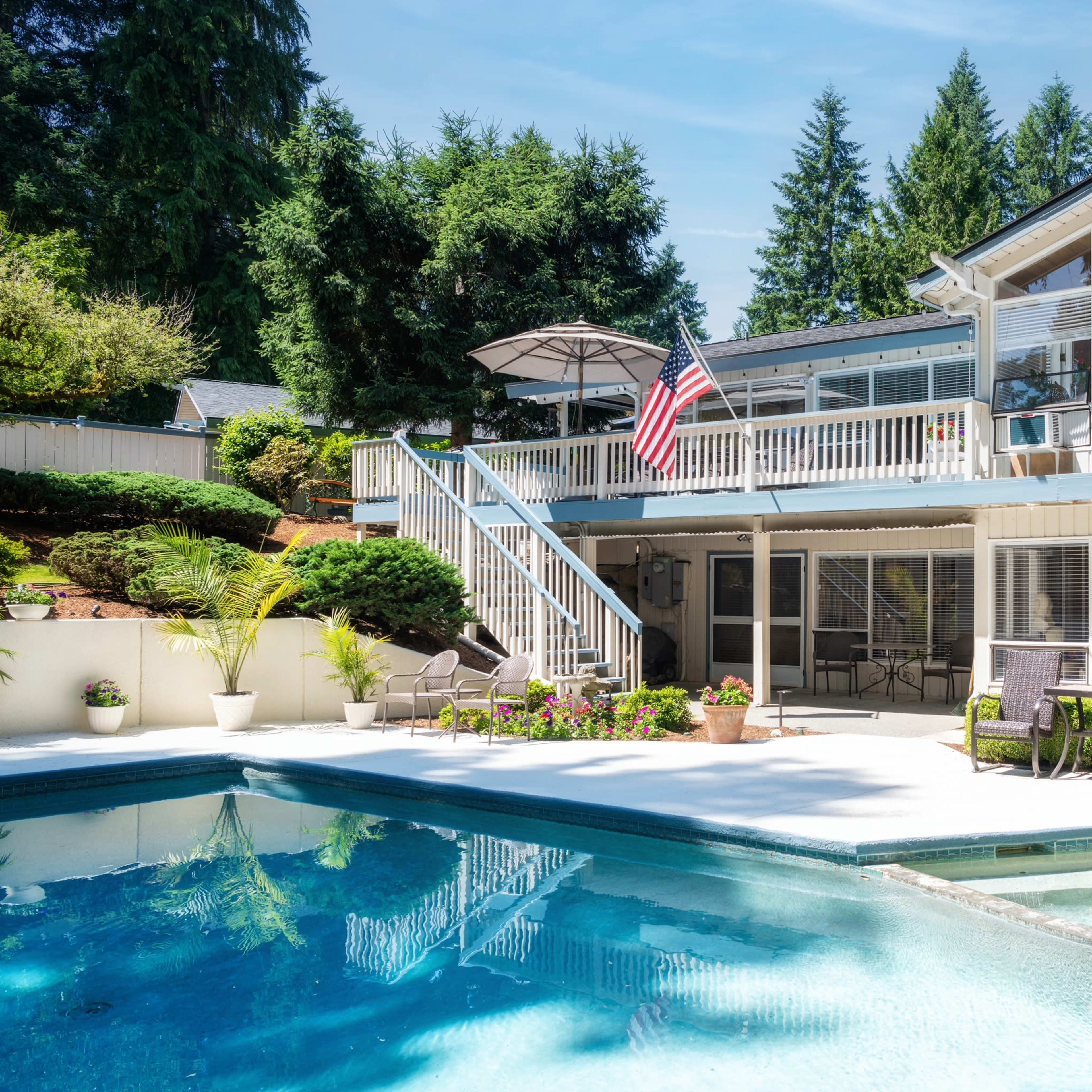 A large pool area is surrounded by a well-maintained patio and a two-story house with an American flag, nestled among trees.