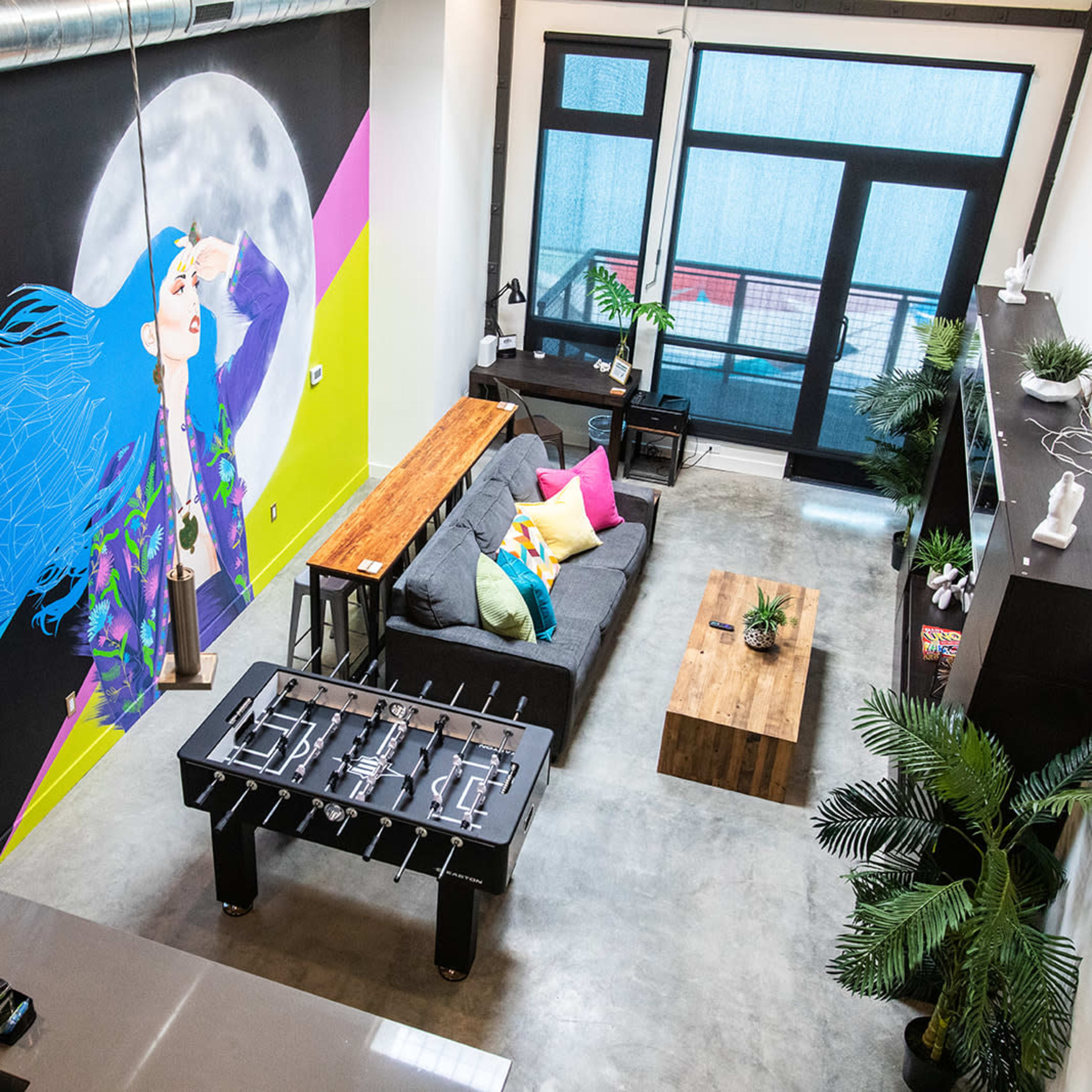A modern living space with a large, colorful mural, a gray sofa with bright cushions, a wooden coffee table, a foosball table, and large windows letting in natural light.