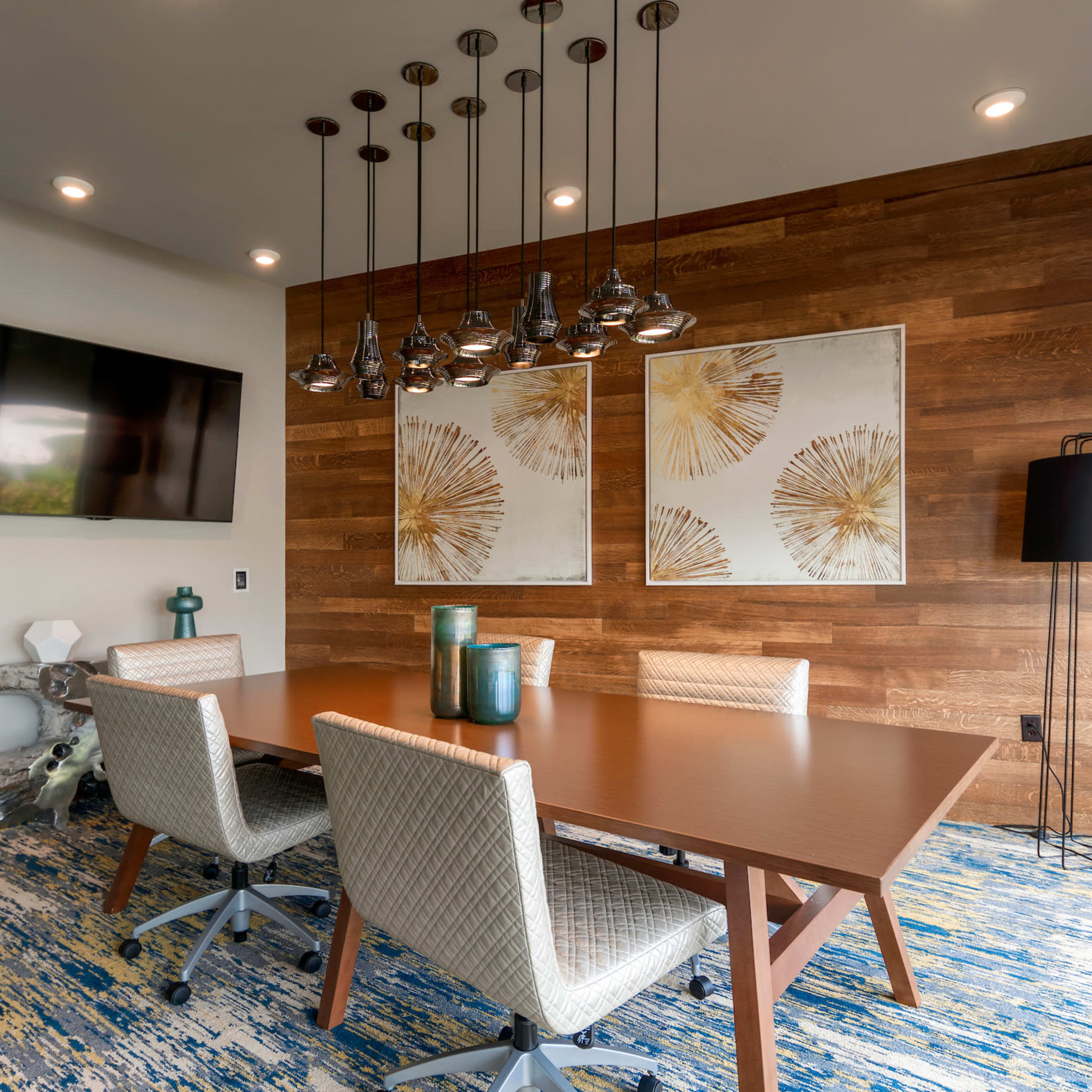 A modern meeting room features a wooden table surrounded by upholstered chairs, with pendant lights hanging above and artwork on the walls.