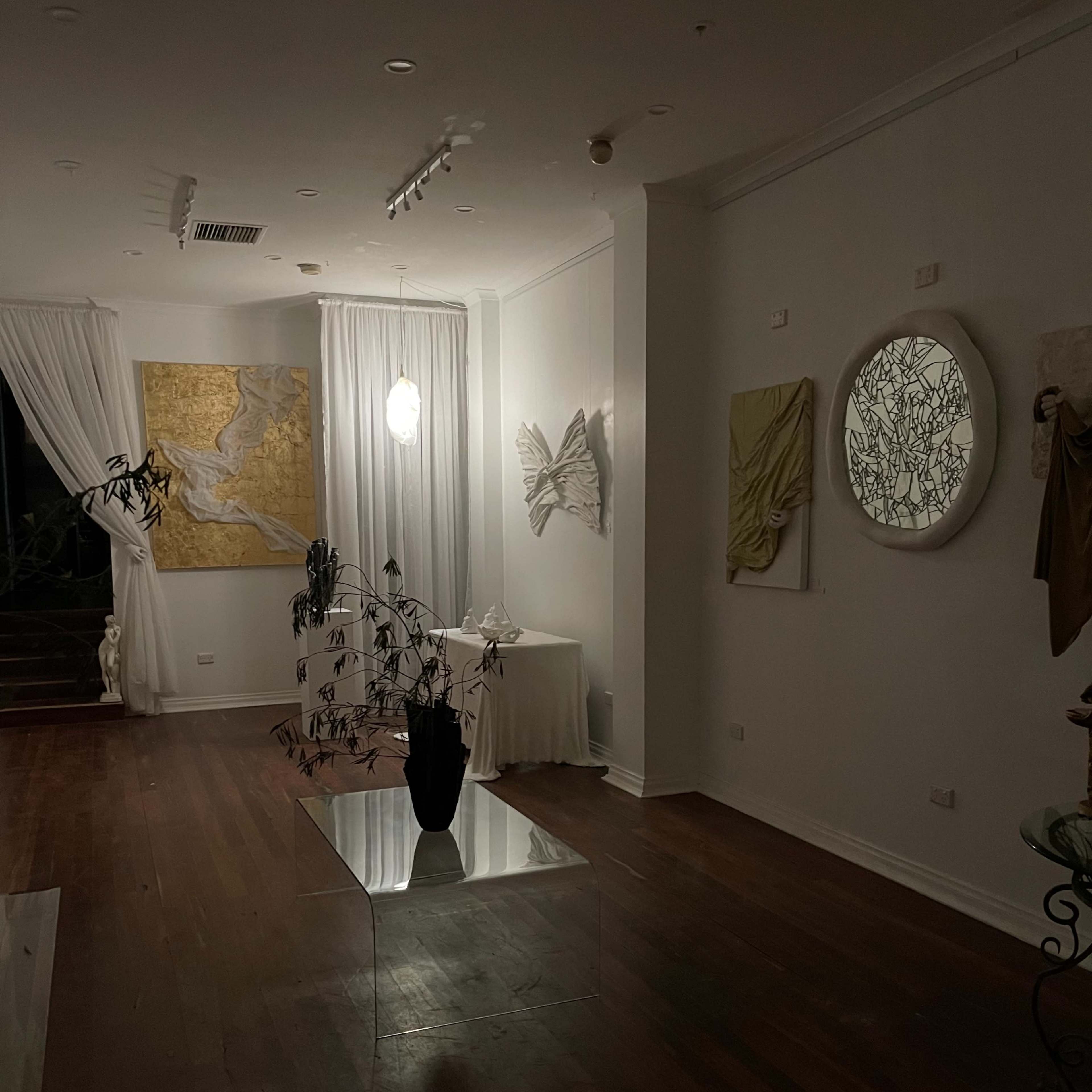 A dimly lit art gallery featuring white walls adorned with abstract artworks and a clear glass table with a plant arrangement.