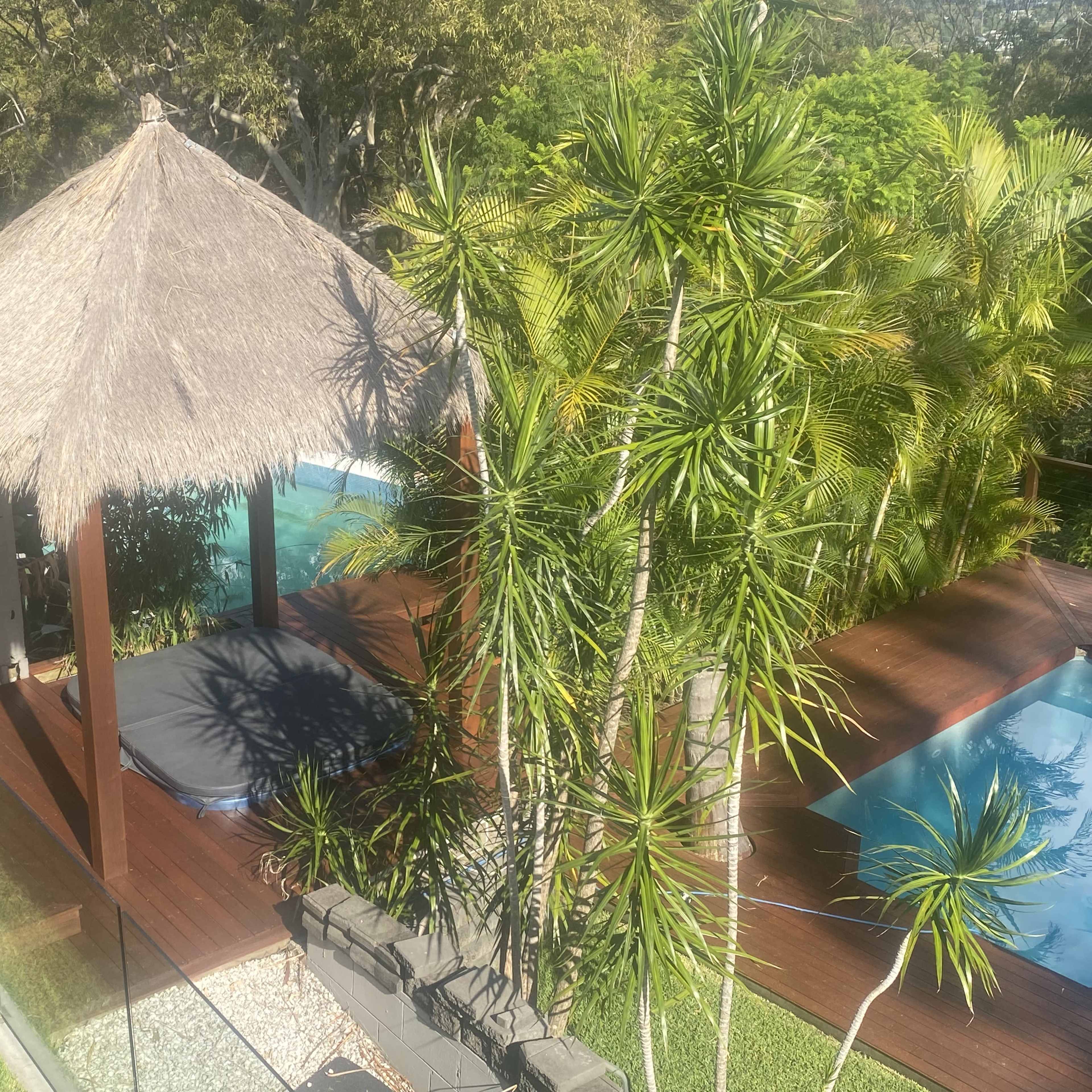 A thatched-roof gazebo sits on a wooden deck near a swimming pool and surrounded by lush greenery.