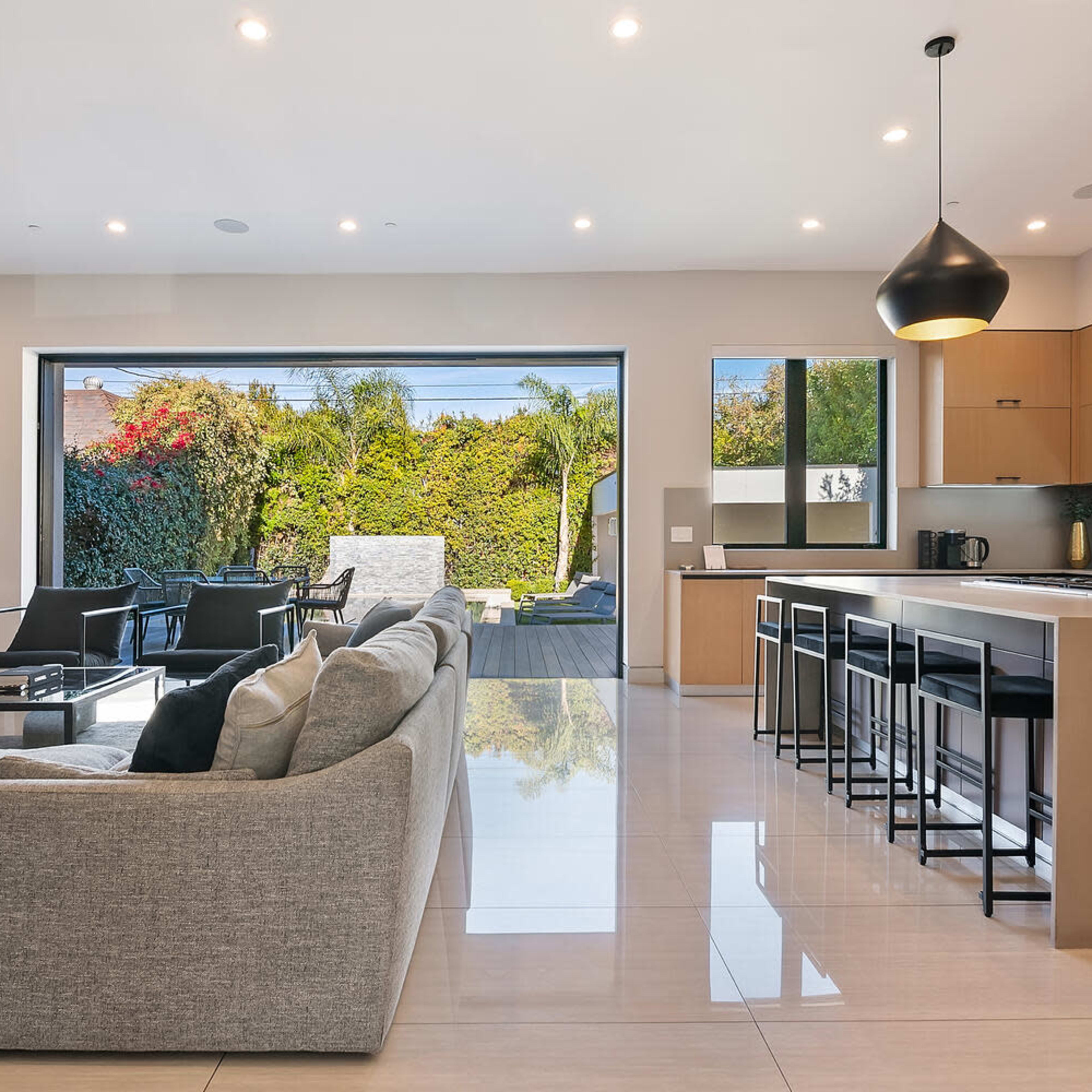 A modern open-concept living area featuring a gray sectional sofa, a kitchen with wooden cabinets, and large glass doors that lead to an outdoor patio.