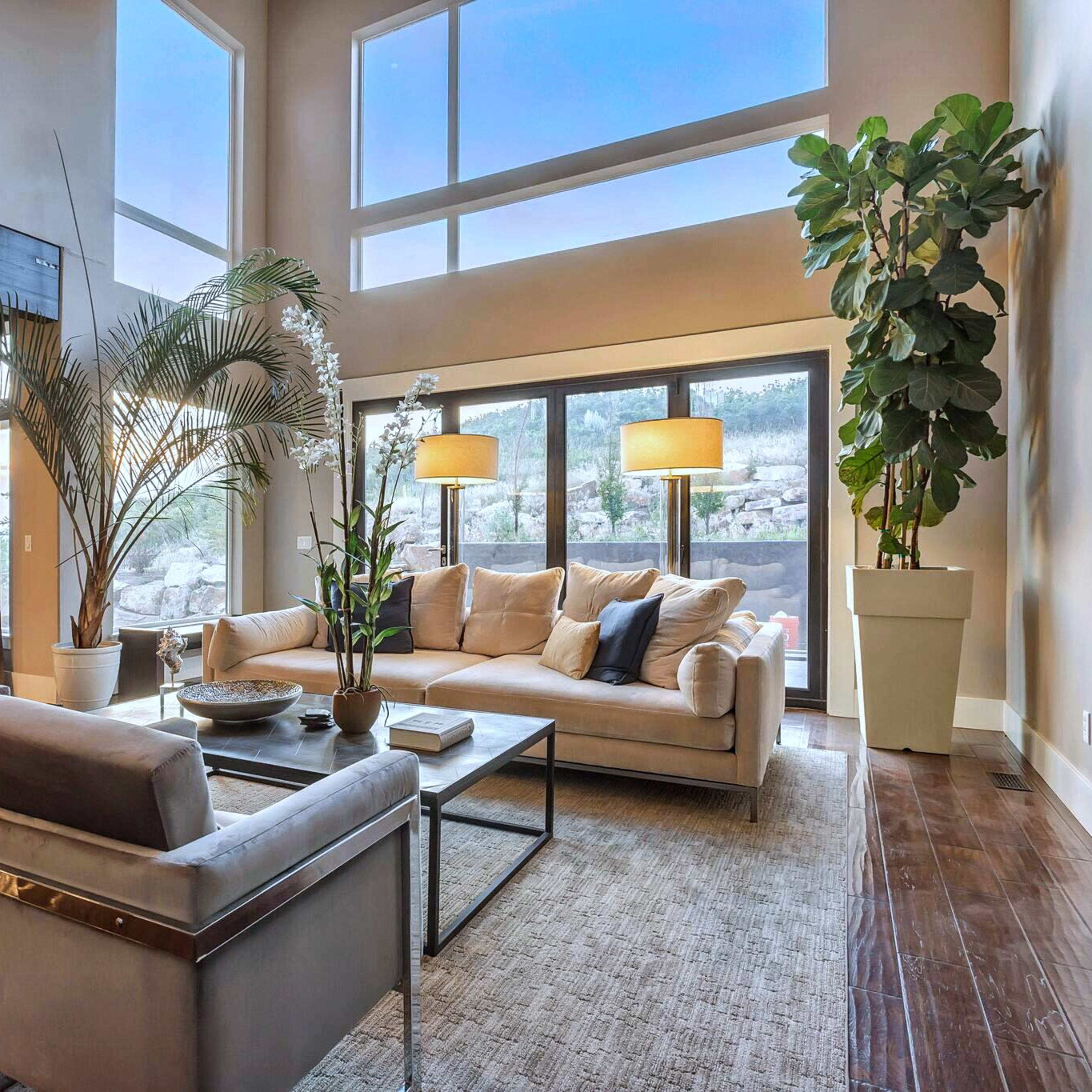 A modern living room with large windows, a light-colored sofa, two armchairs, and various indoor plants.