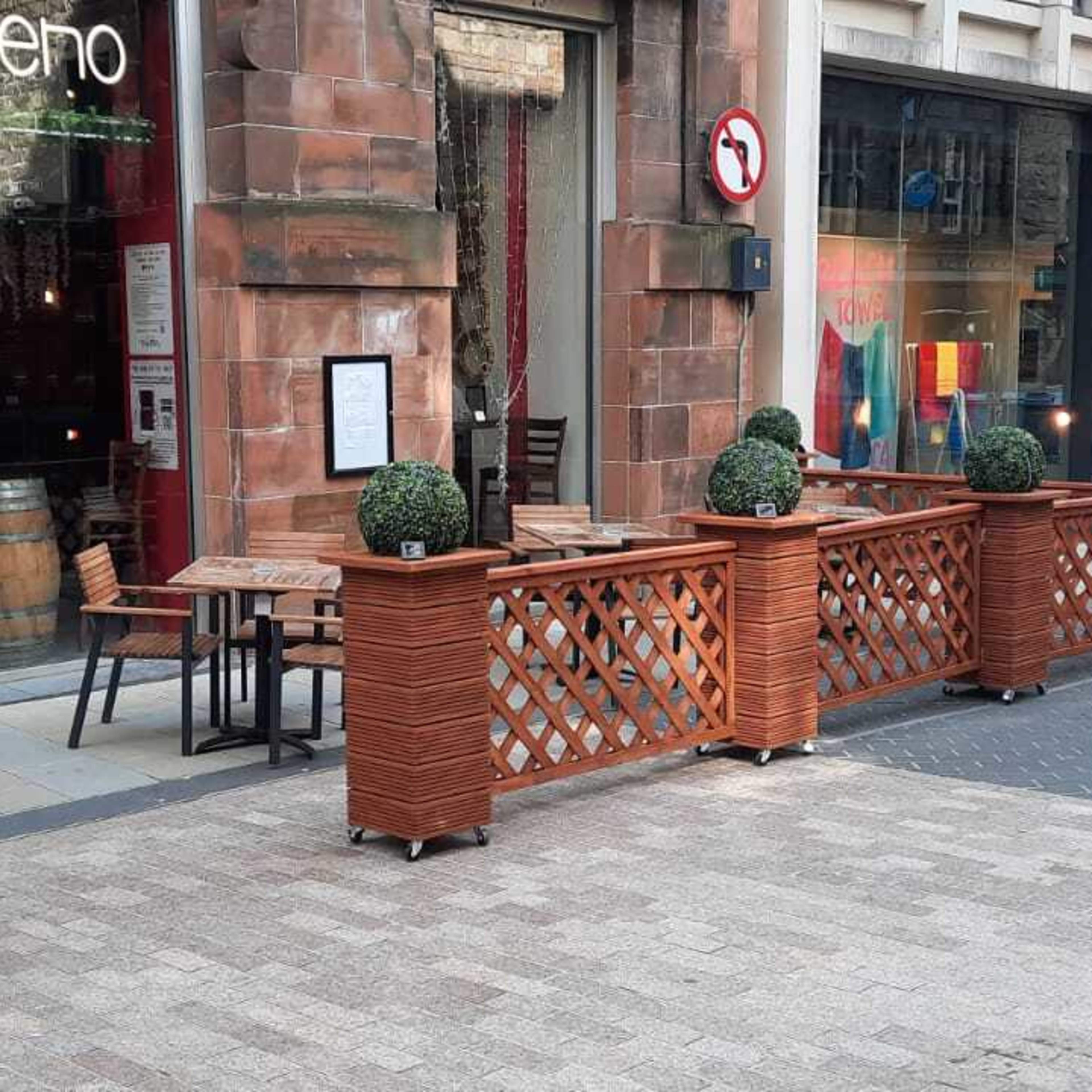 An outdoor seating area with wooden barriers and potted topiaries in front of a restaurant called "Veenoo" on a street with pedestrians walking by.