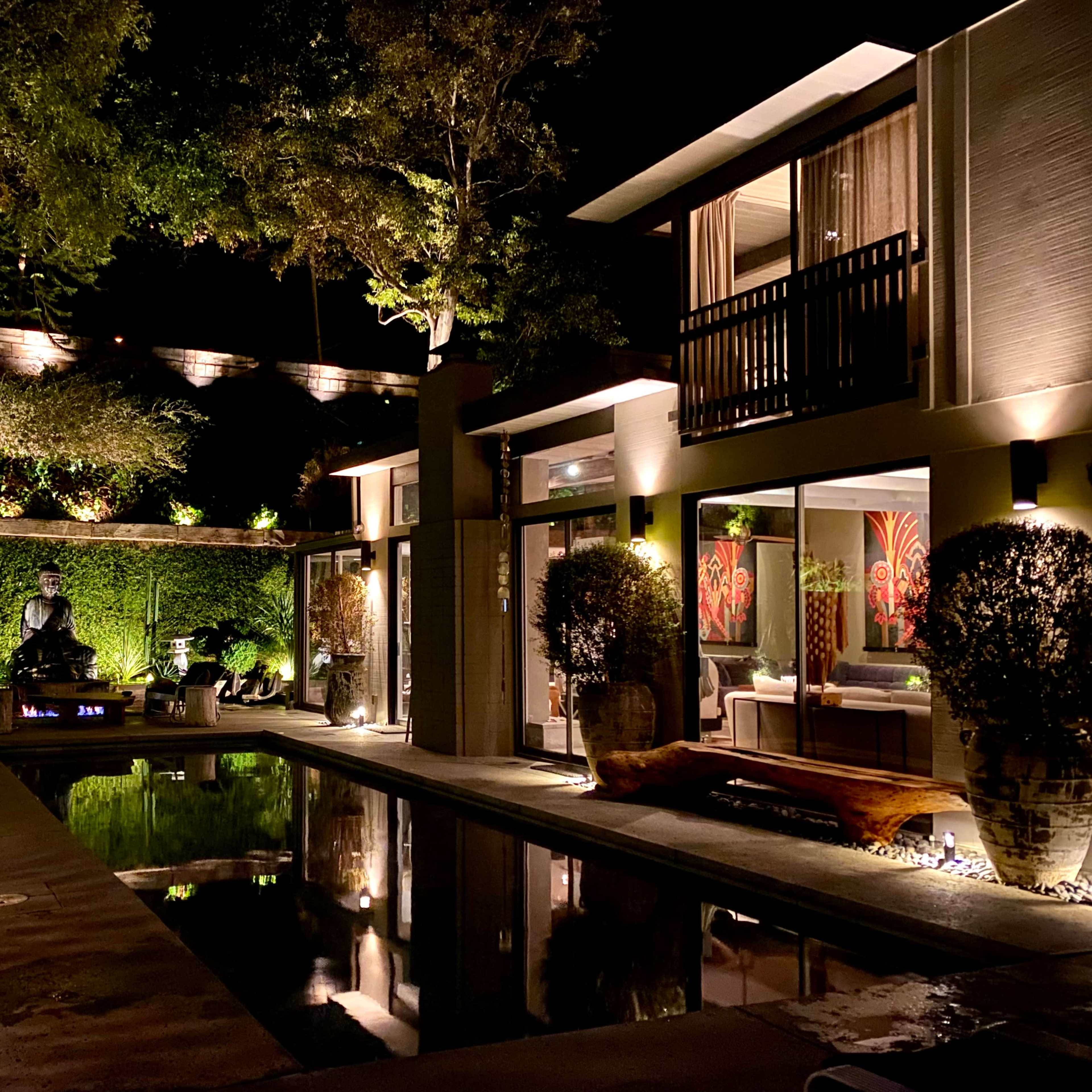 A modern outdoor pool area at night, featuring landscaped greenery, illuminated trees, and a statue surrounded by decorative elements.