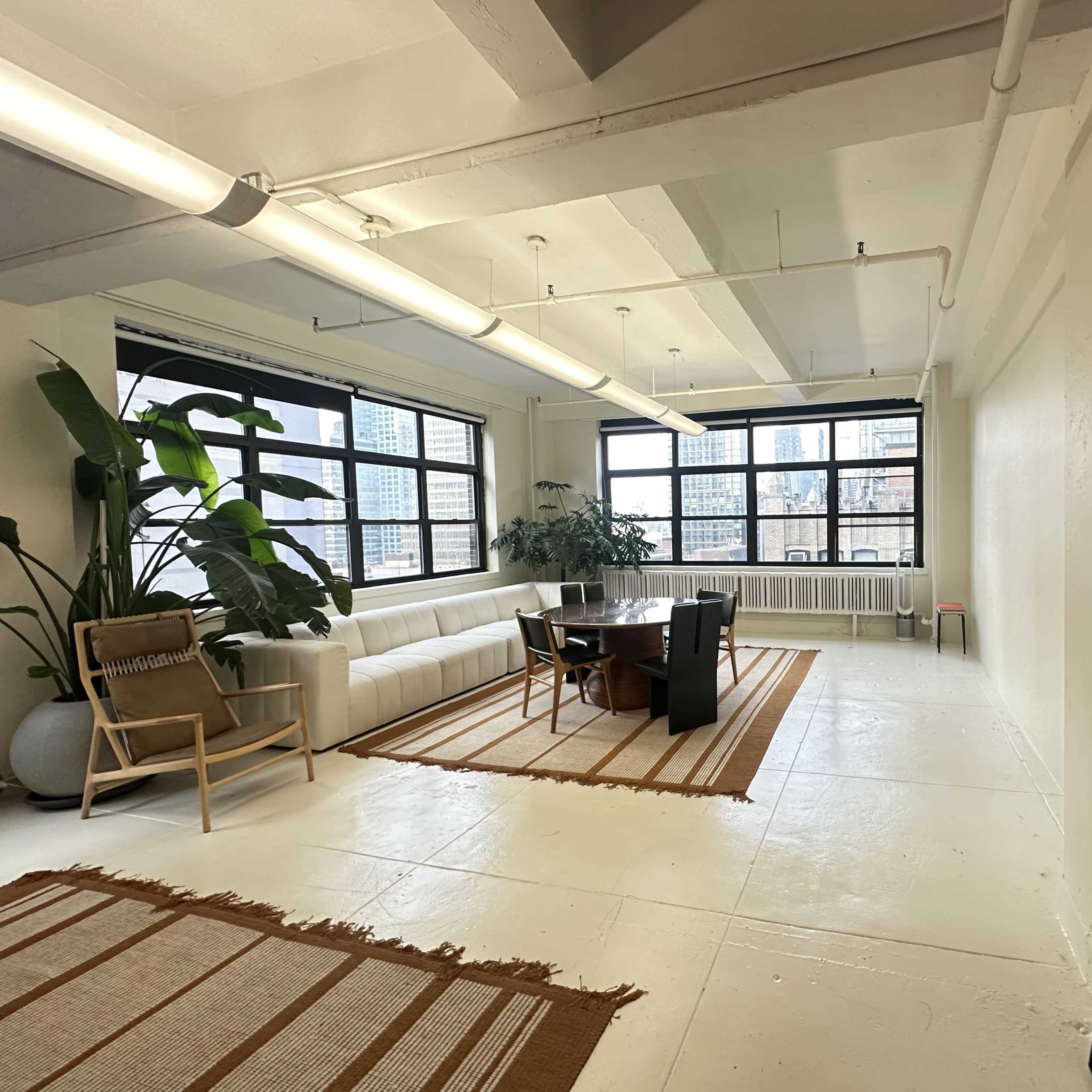 A spacious interior room with large windows, featuring a light-colored sofa, a dining table, plants, and a large mirror.