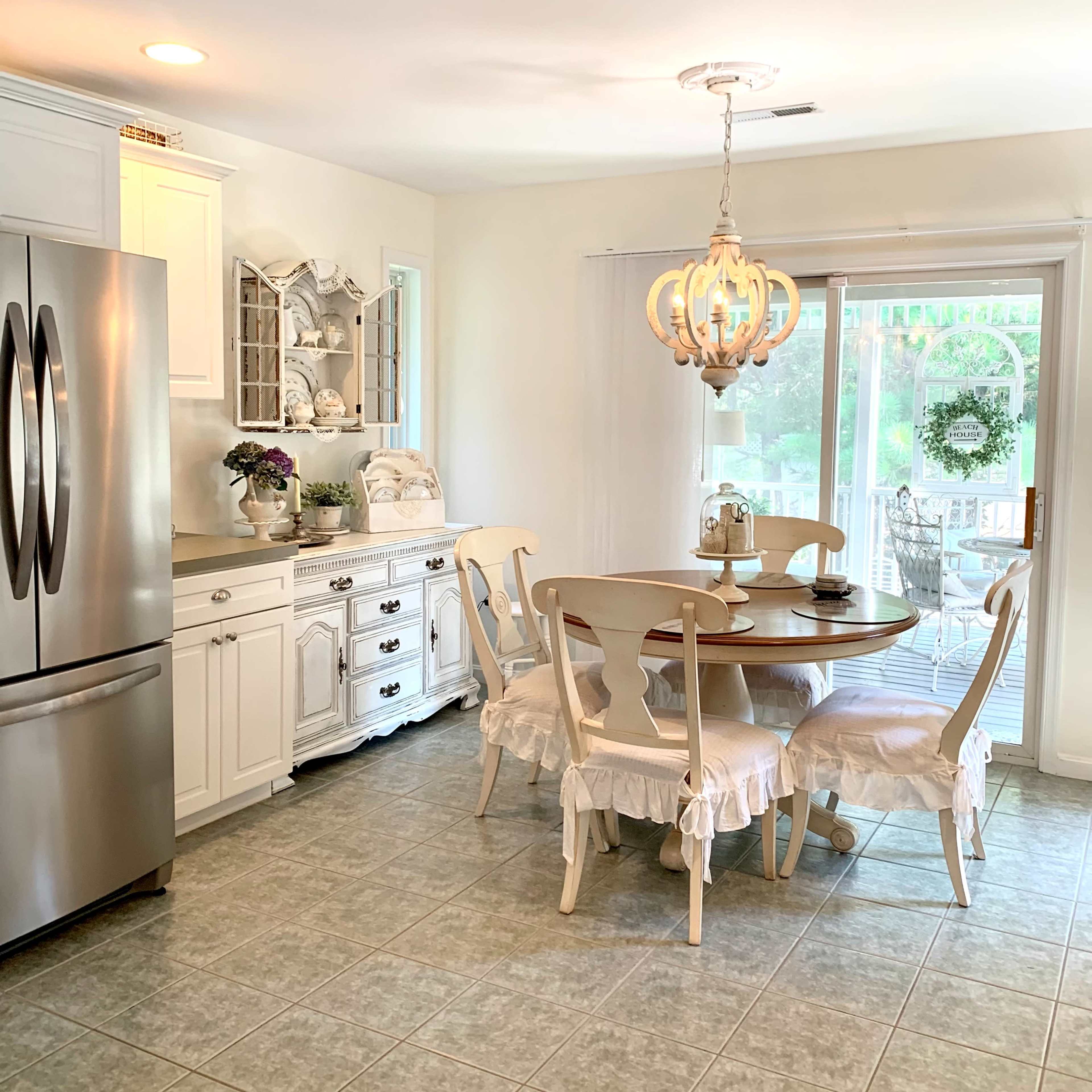 A modern kitchen with a round dining table, four chairs, a stainless steel refrigerator, and a large window leading to a deck.