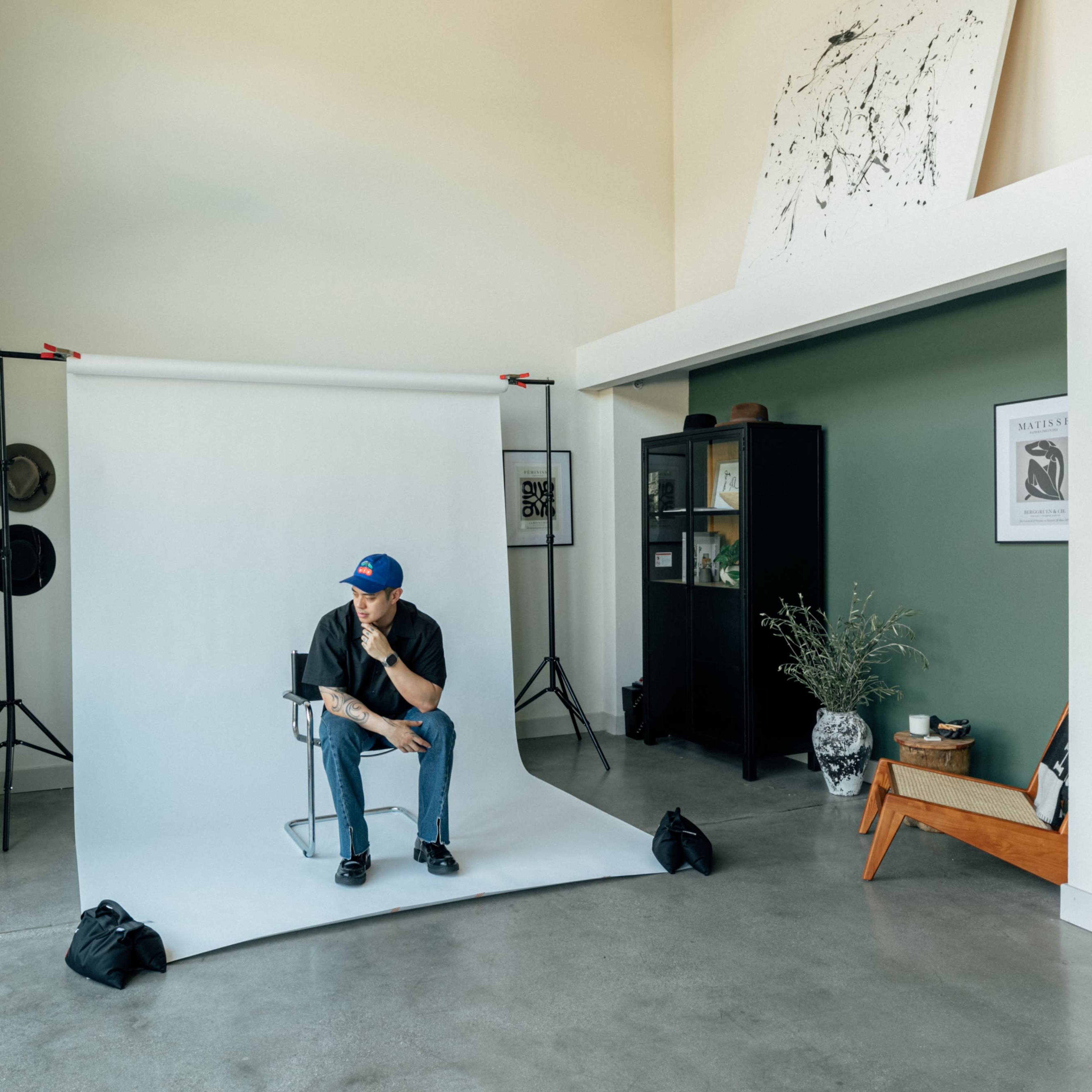 A person sits on a chair in a photography studio with a white backdrop and various decorative elements around the room.