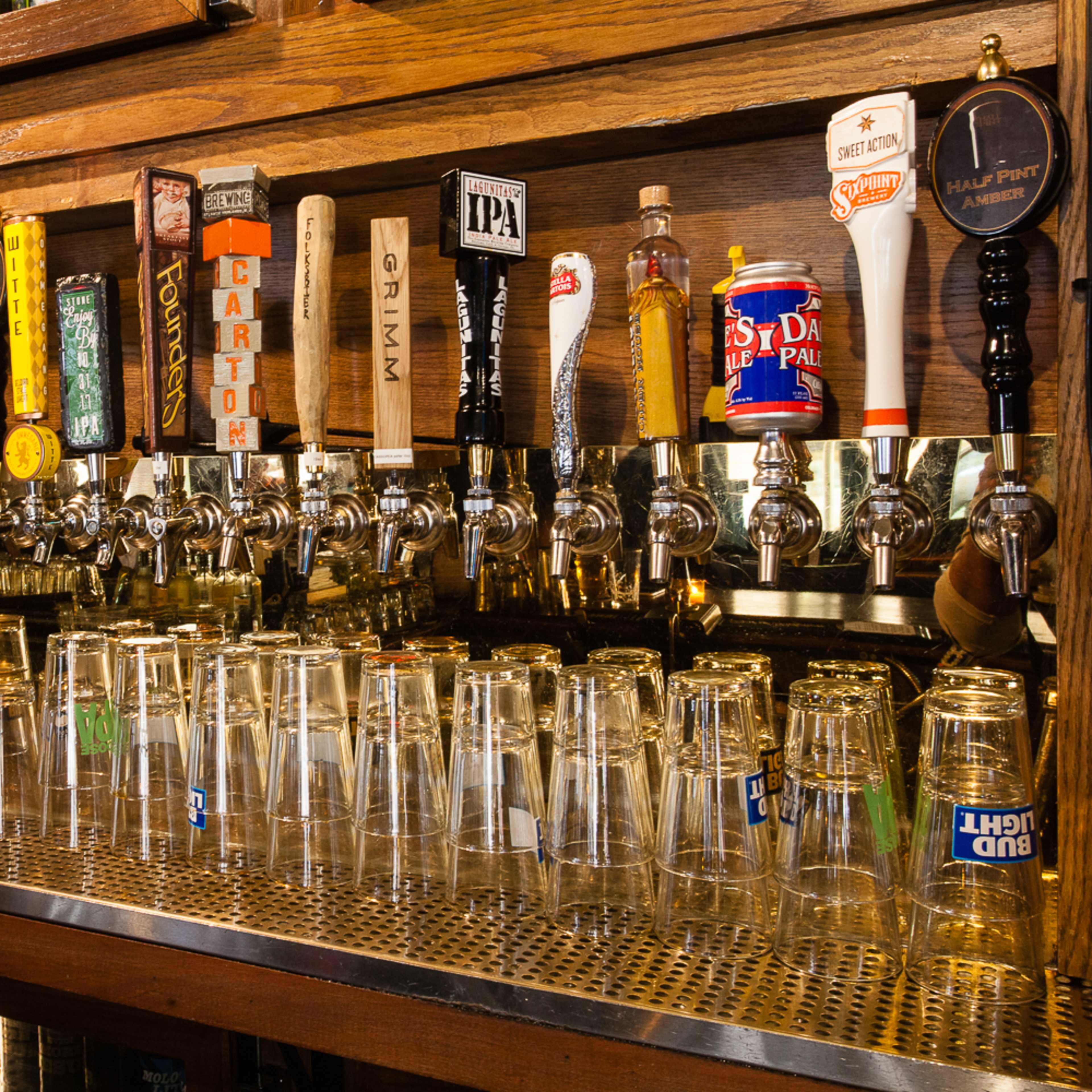 A wooden bar with multiple beer taps and a row of glass mugs lined up on a metal surface.