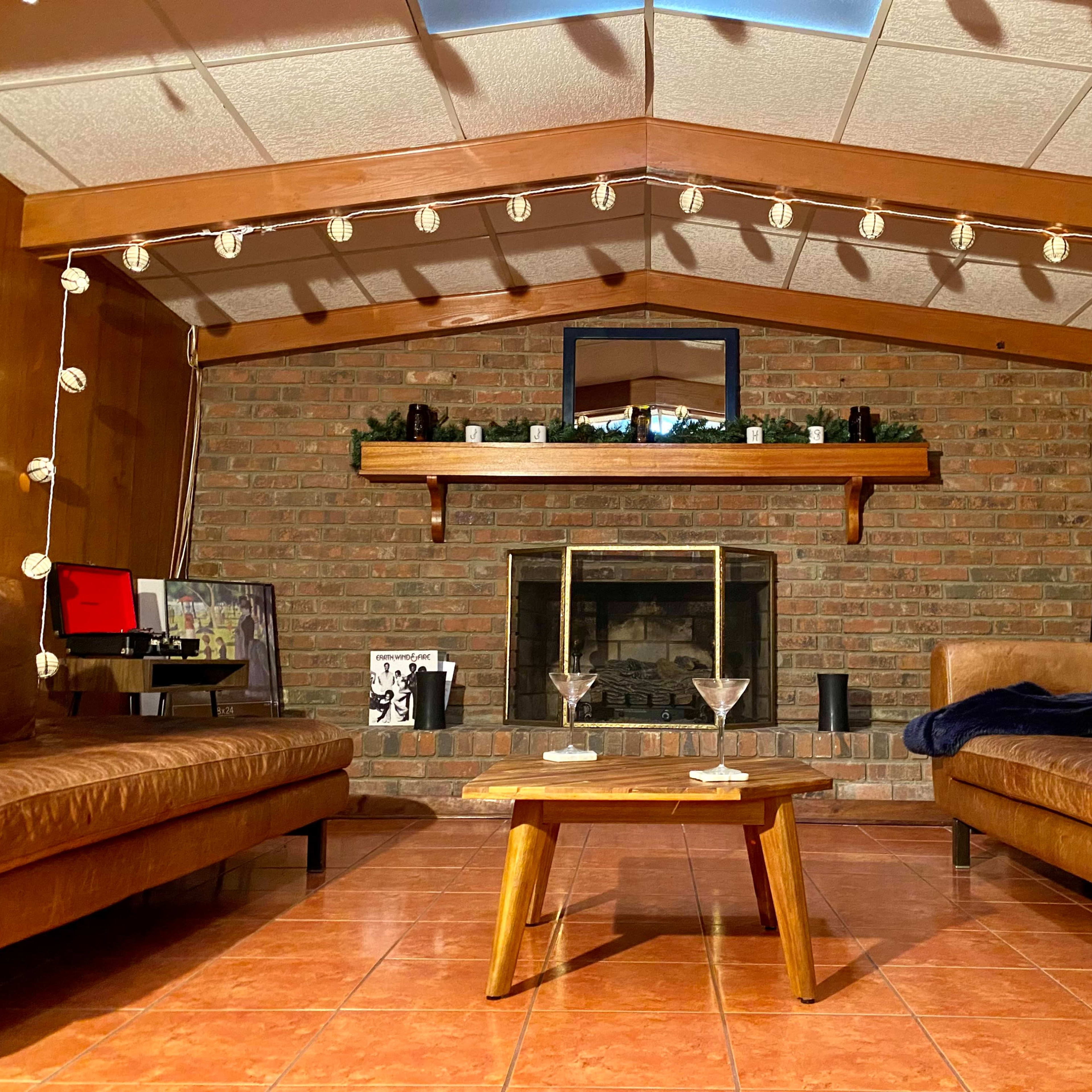 A cozy living room features two leather couches facing a brick fireplace, with a wooden coffee table in the center and string lights overhead.
