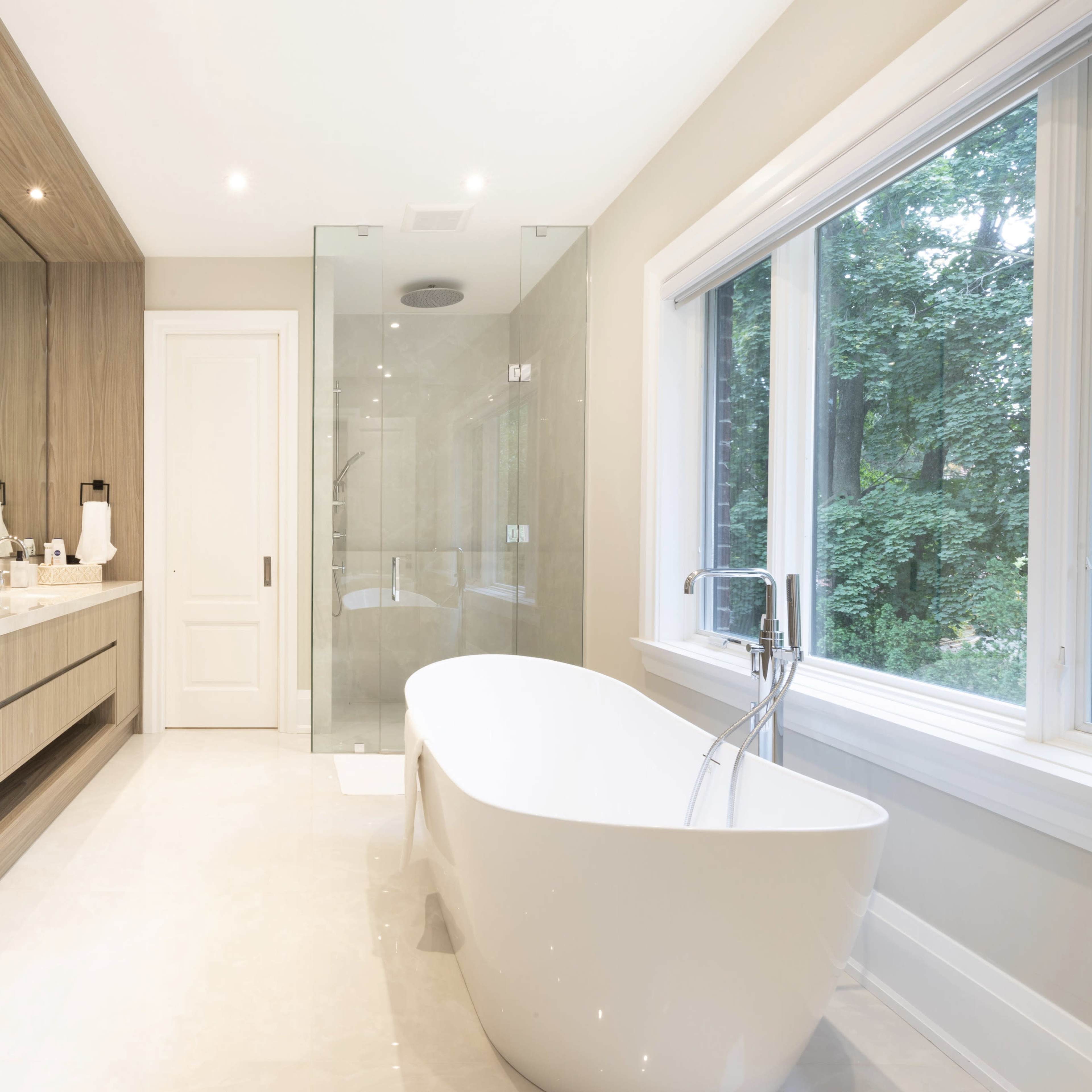 A modern bathroom featuring a freestanding bathtub, a double vanity with wooden accents, and a glass shower enclosure beside a large window.