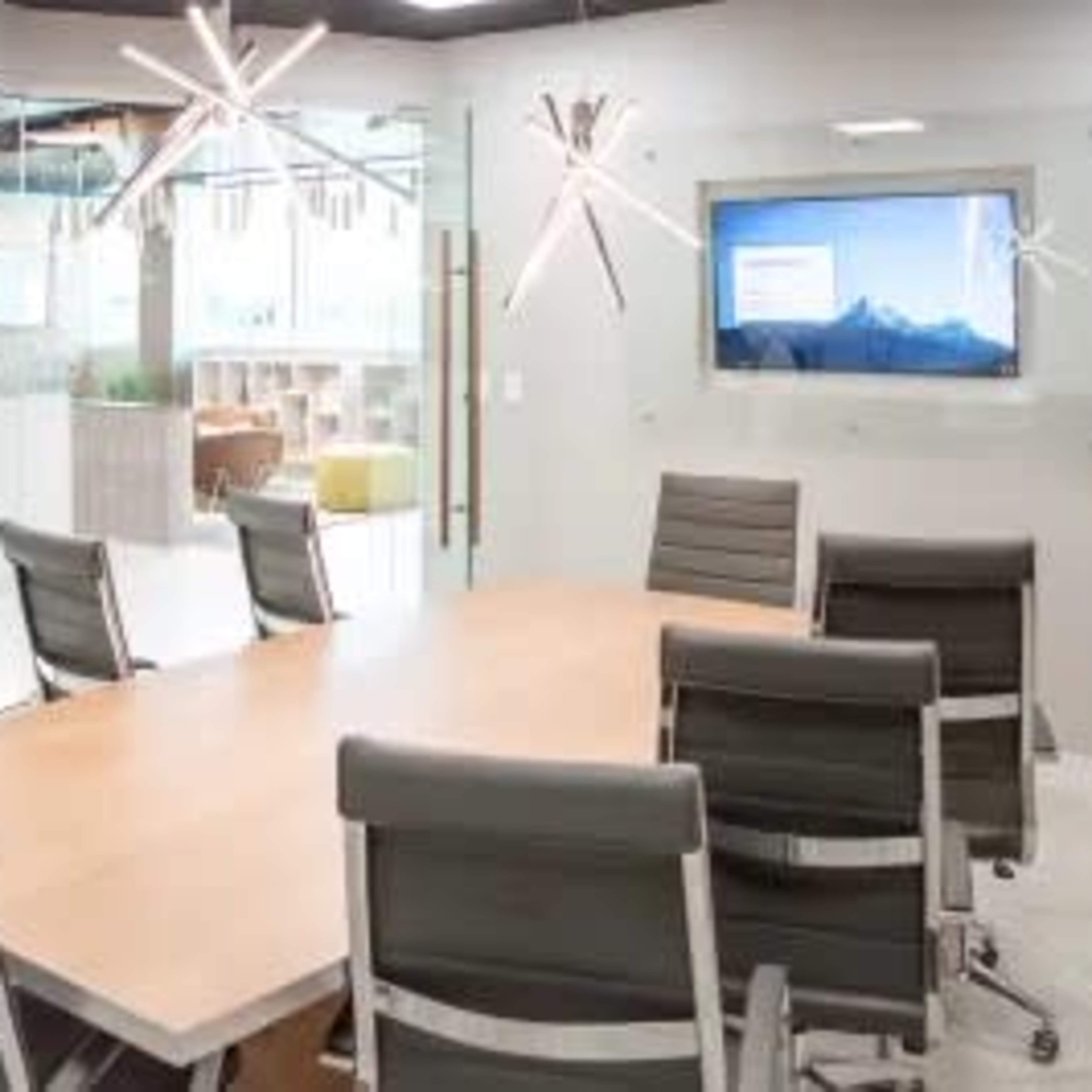A modern conference room features a long wooden table surrounded by ergonomic chairs, with a large screen mounted on the wall and stylish light fixtures overhead.