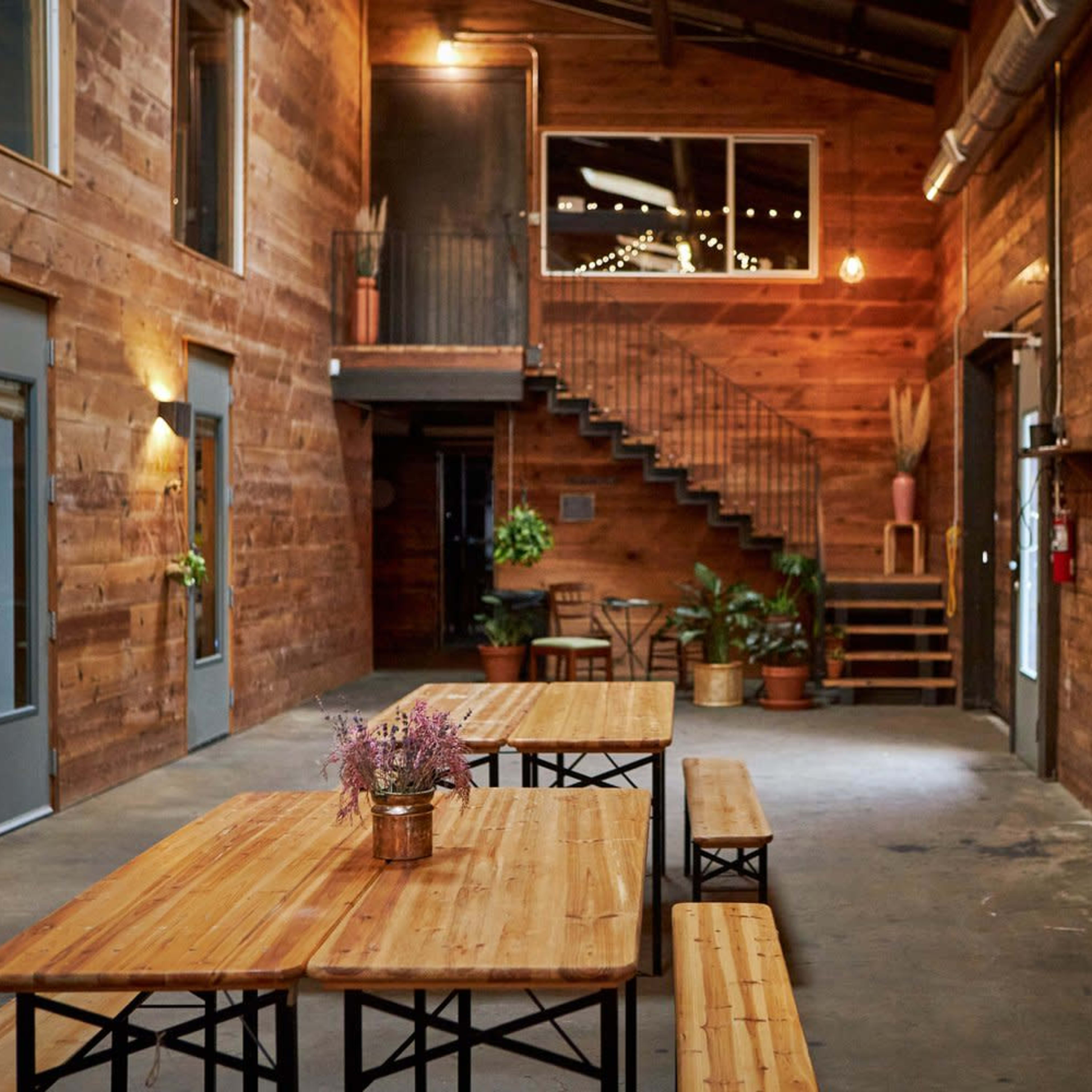 A wooden interior space with multiple tables arranged along the floor, a staircase leading to an upper level, and potted plants along the walls.
