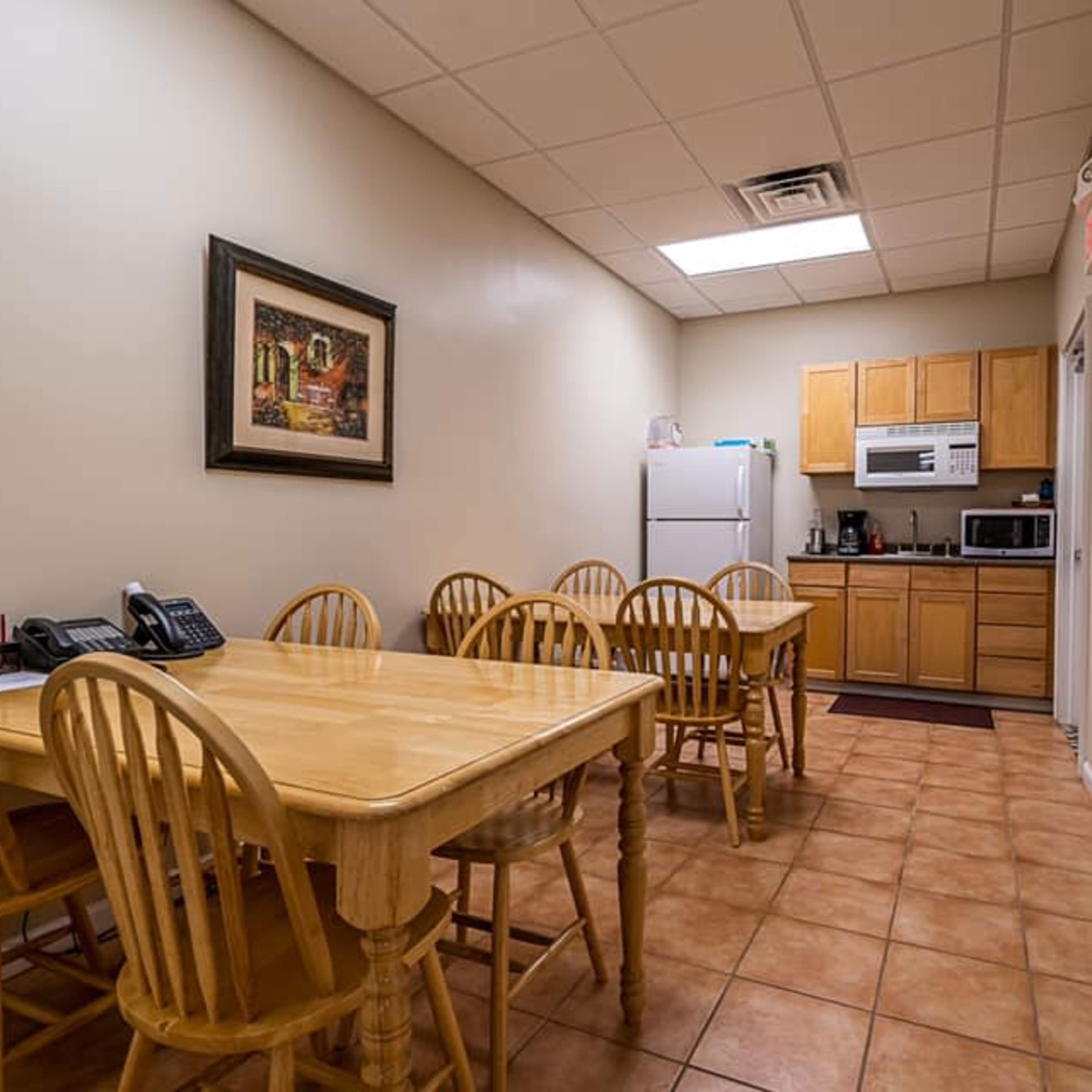 A small kitchen area features two tables with wooden chairs, a whiteboard, and kitchen appliances including a refrigerator and microwave on one side.