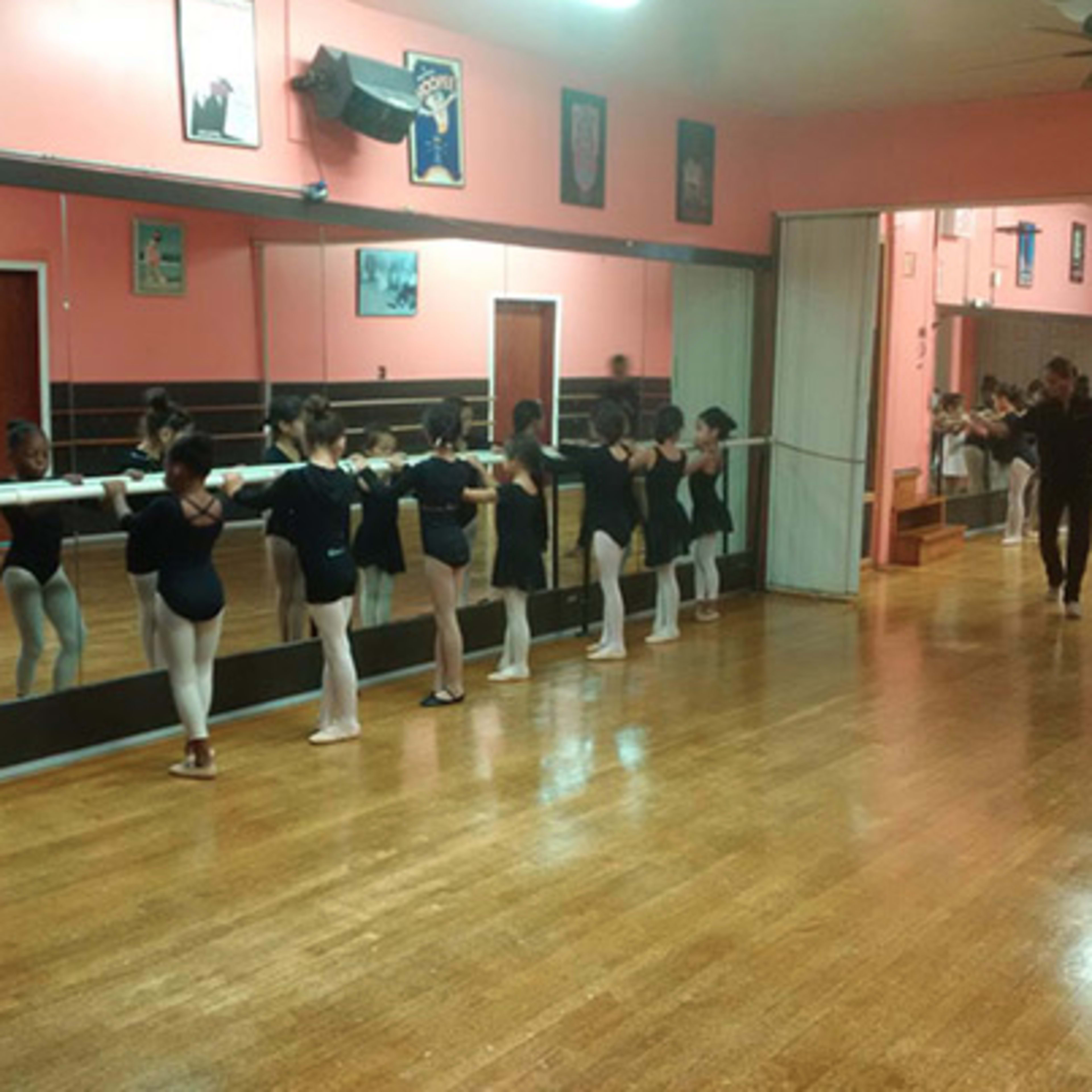A dance studio features young ballet students in black leotards practicing at a mirrored barre under warm lighting.