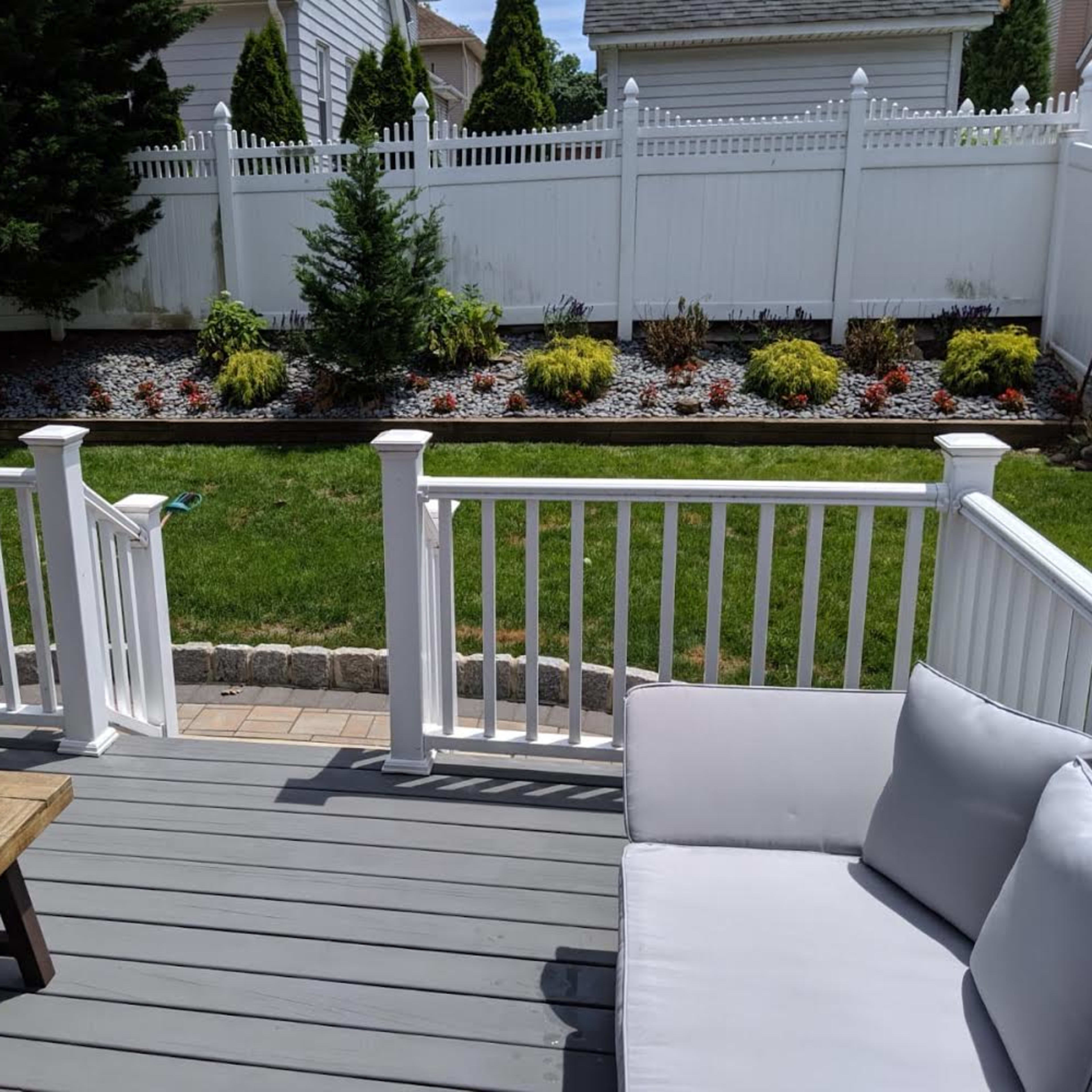 A backyard deck with a wooden table and white sofa, overlooking a landscaped garden with a white fence and colorful flower beds.