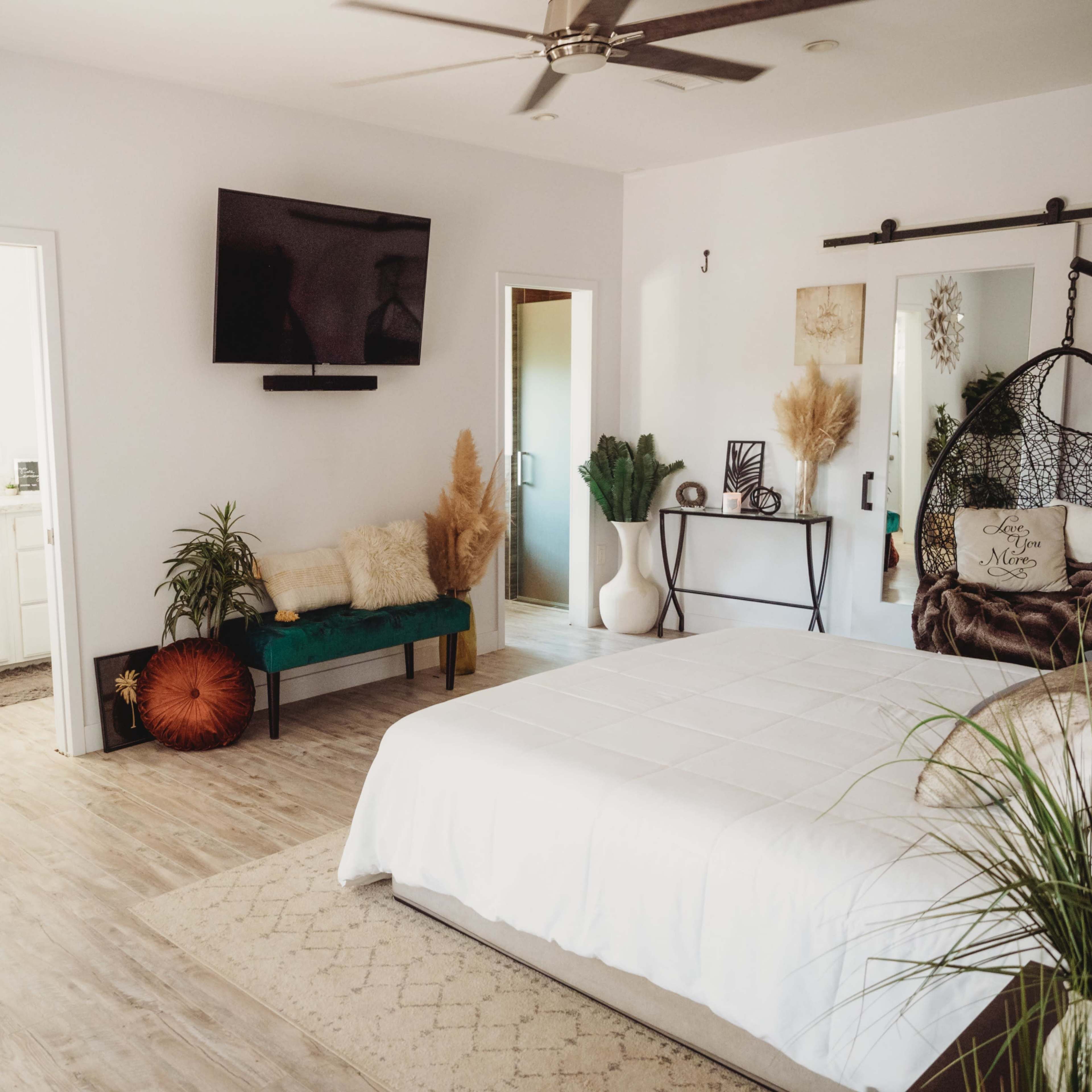 A spacious bedroom featuring a white bed, a green bench, a television mounted on the wall, and a bathroom accessible through an adjoining door.