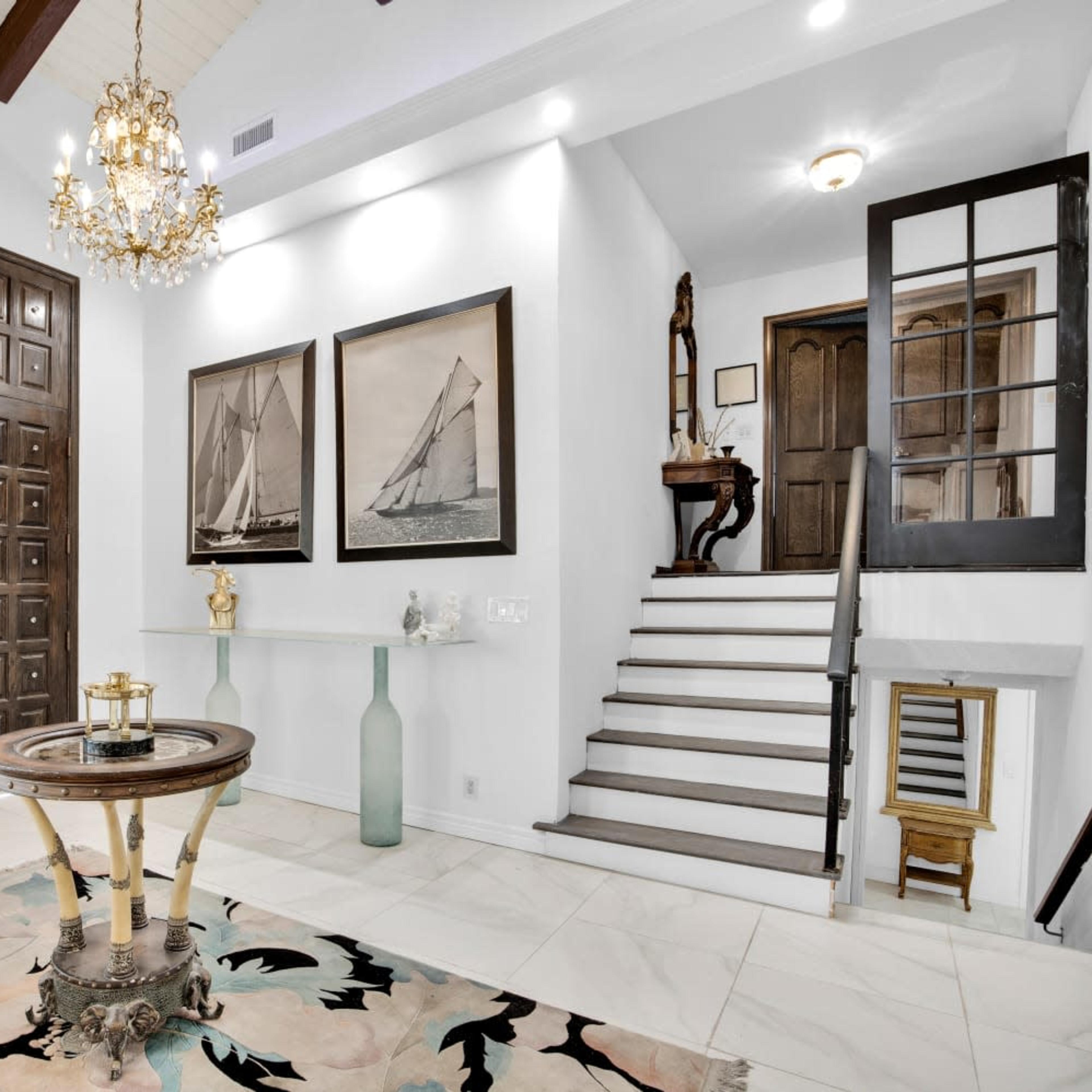 A spacious entryway featuring a large wooden door, decorative chandeliers, framed pictures of sailboats, and a staircase leading to an upper level.