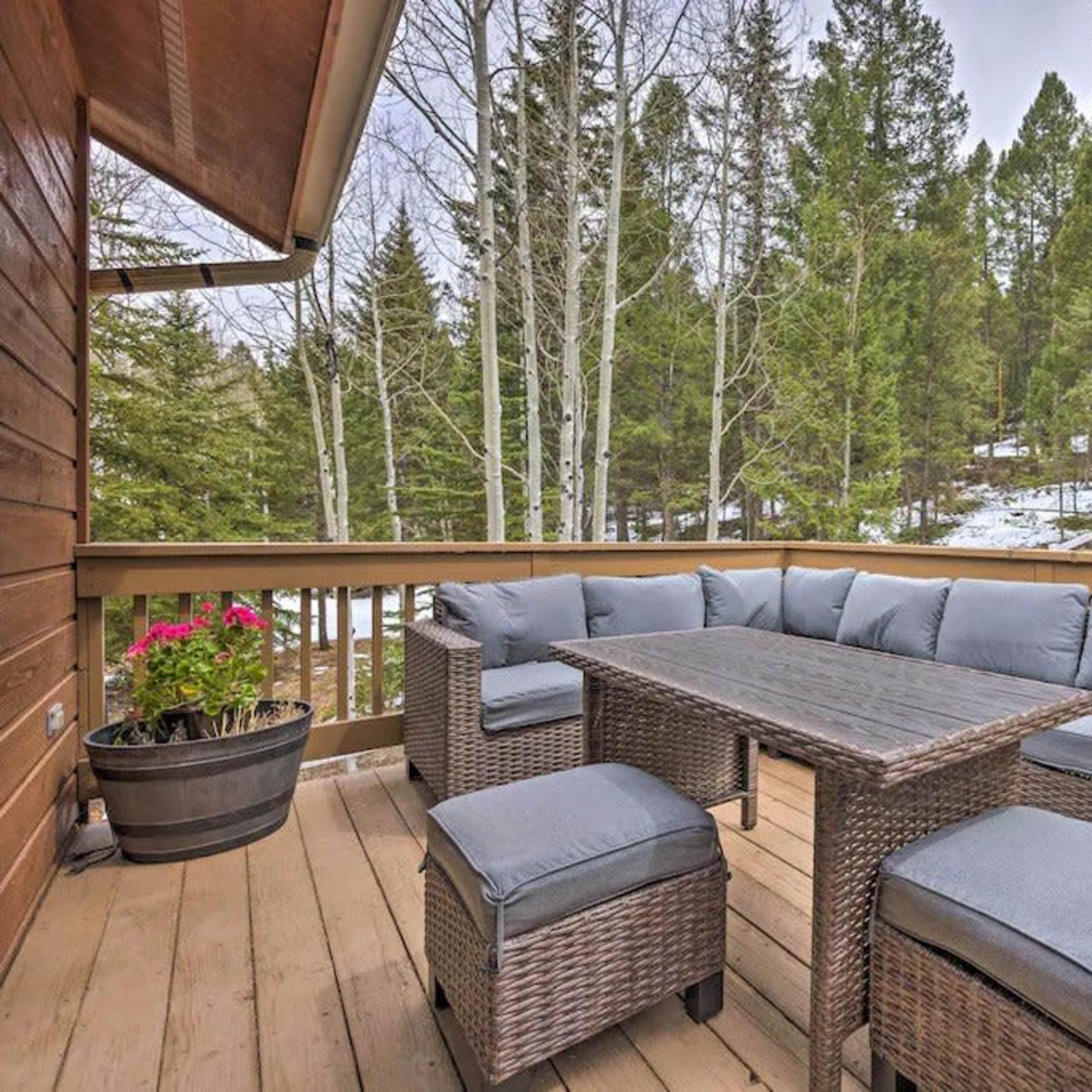 A wooden deck features a rectangular table surrounded by cushioned seating, with planters and a backdrop of tall pine trees and a hint of snow.