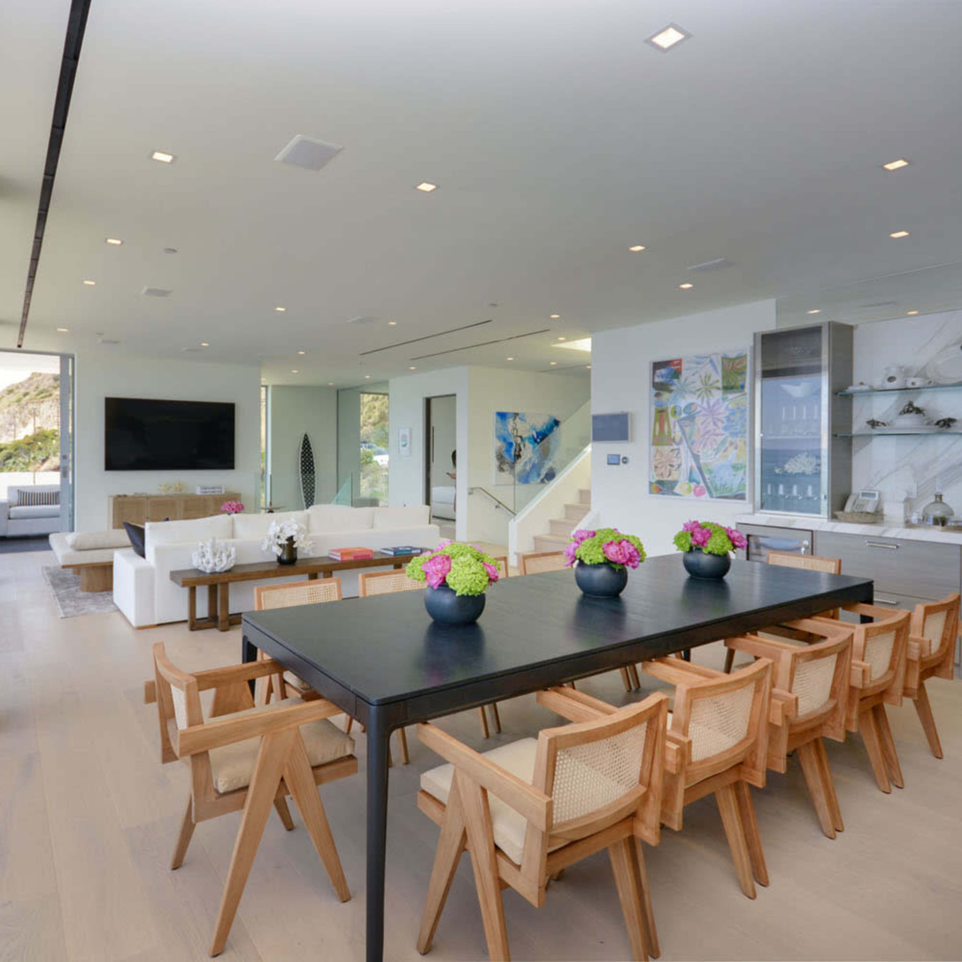 A modern dining area with a long table surrounded by wooden chairs, adjacent to a sleek kitchen and a living space with large windows overlooking a view.