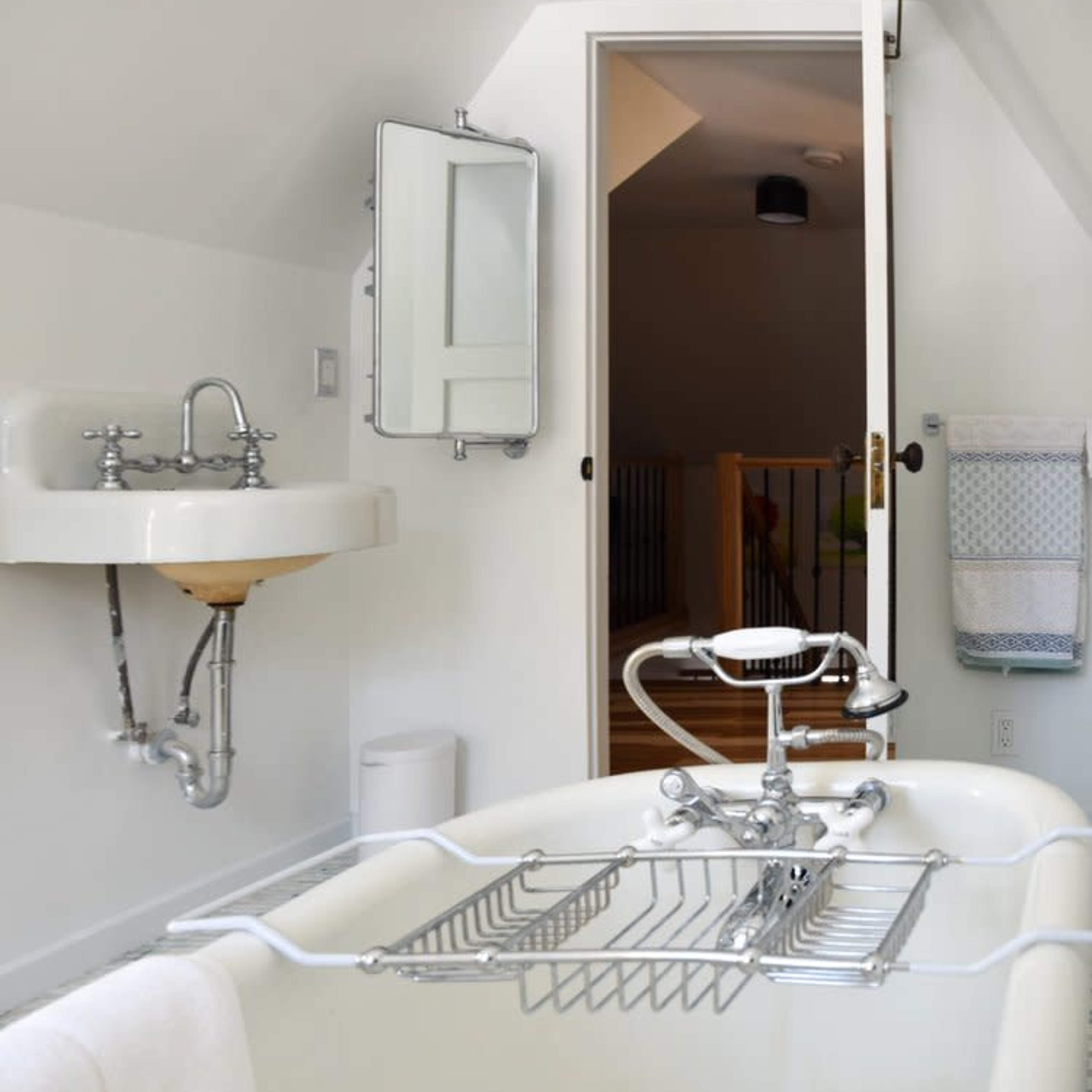 A bathroom featuring a clawfoot bathtub with a metal rack, a wall-mounted sink, and a doorway leading to a staircase.