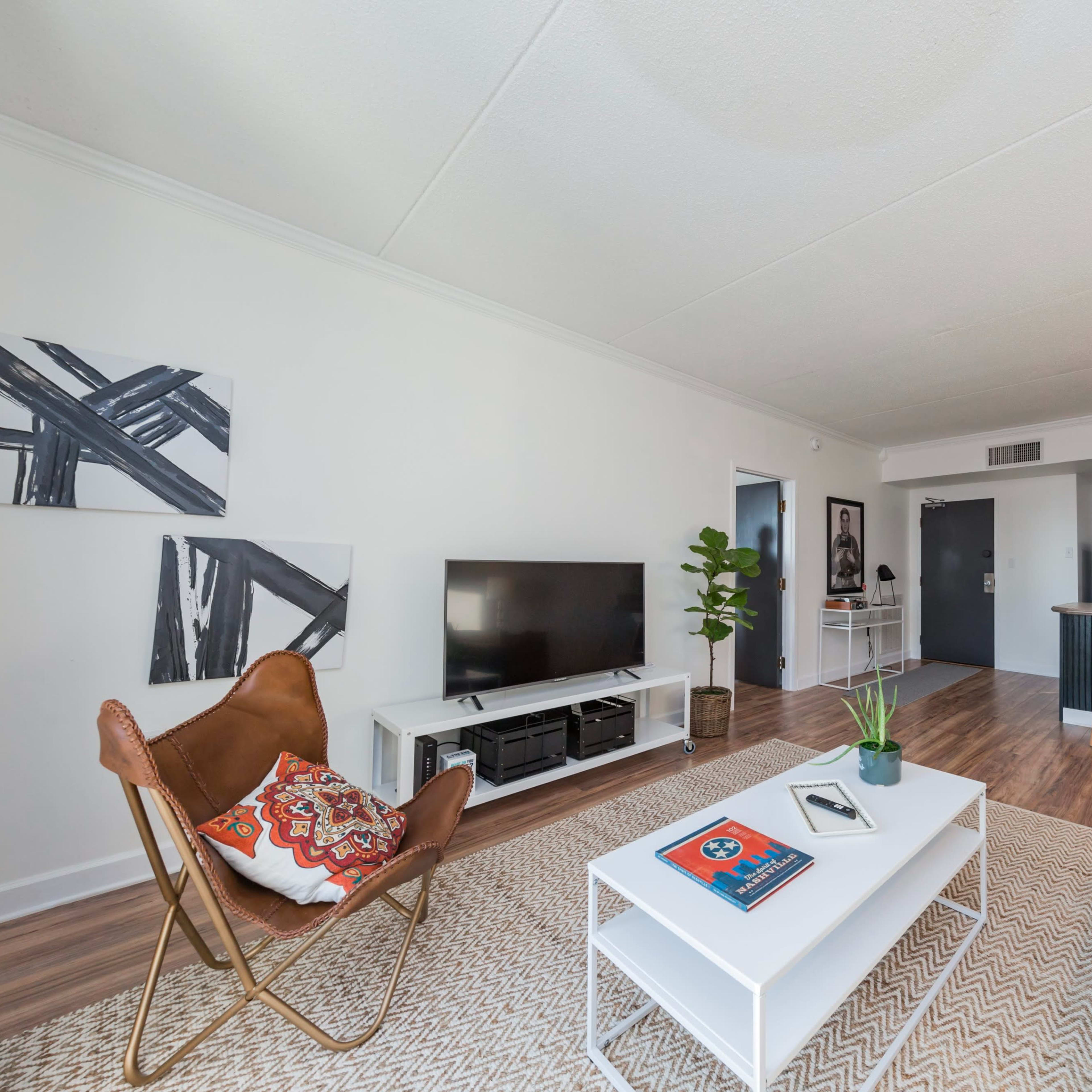 A modern living room with a television, a coffee table, and a combination of seating options including a chair and a sofa, leading into a kitchen area in the background.