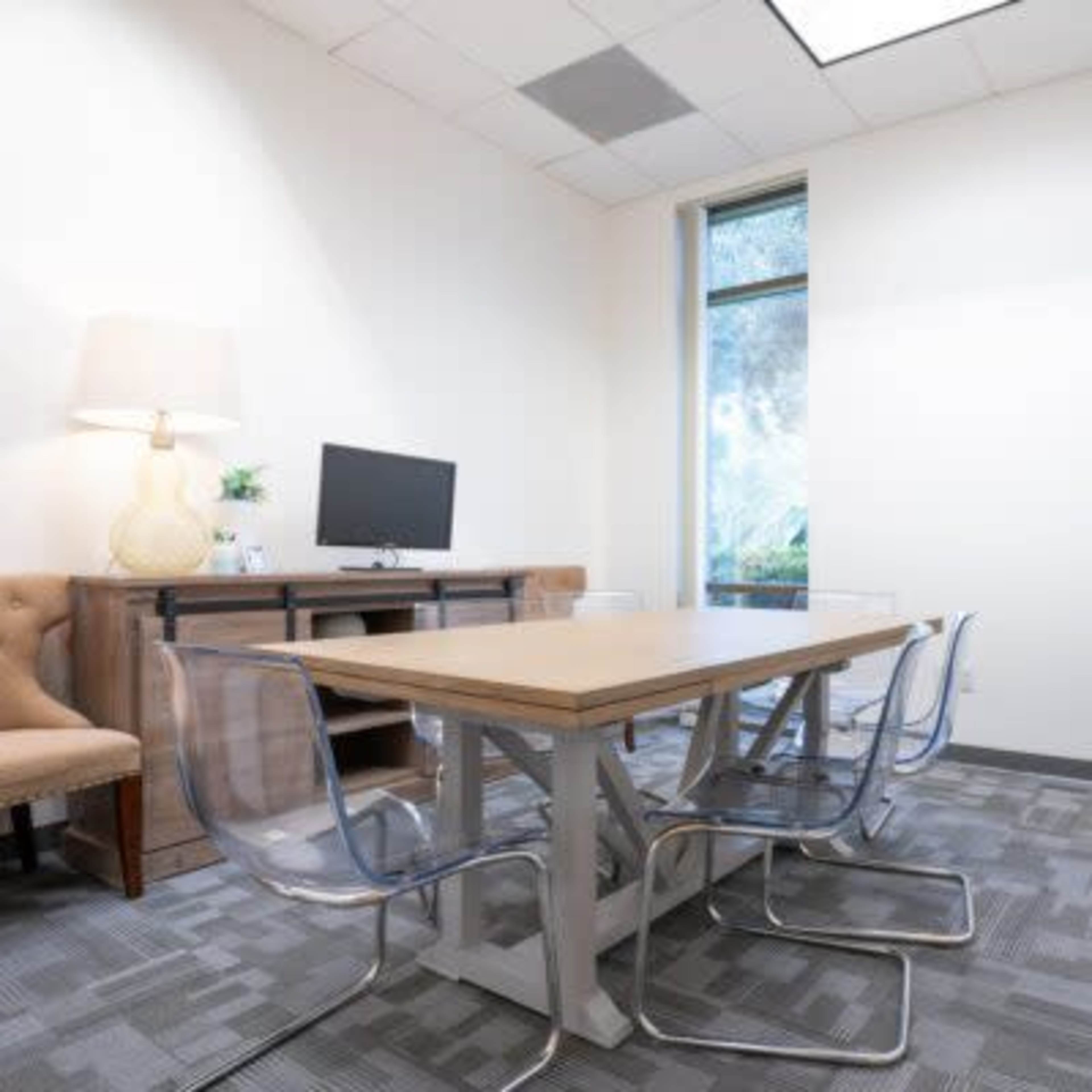 A small meeting room with a wooden table surrounded by clear chairs, a wall-mounted monitor, a lamp, a plant, and a large window.