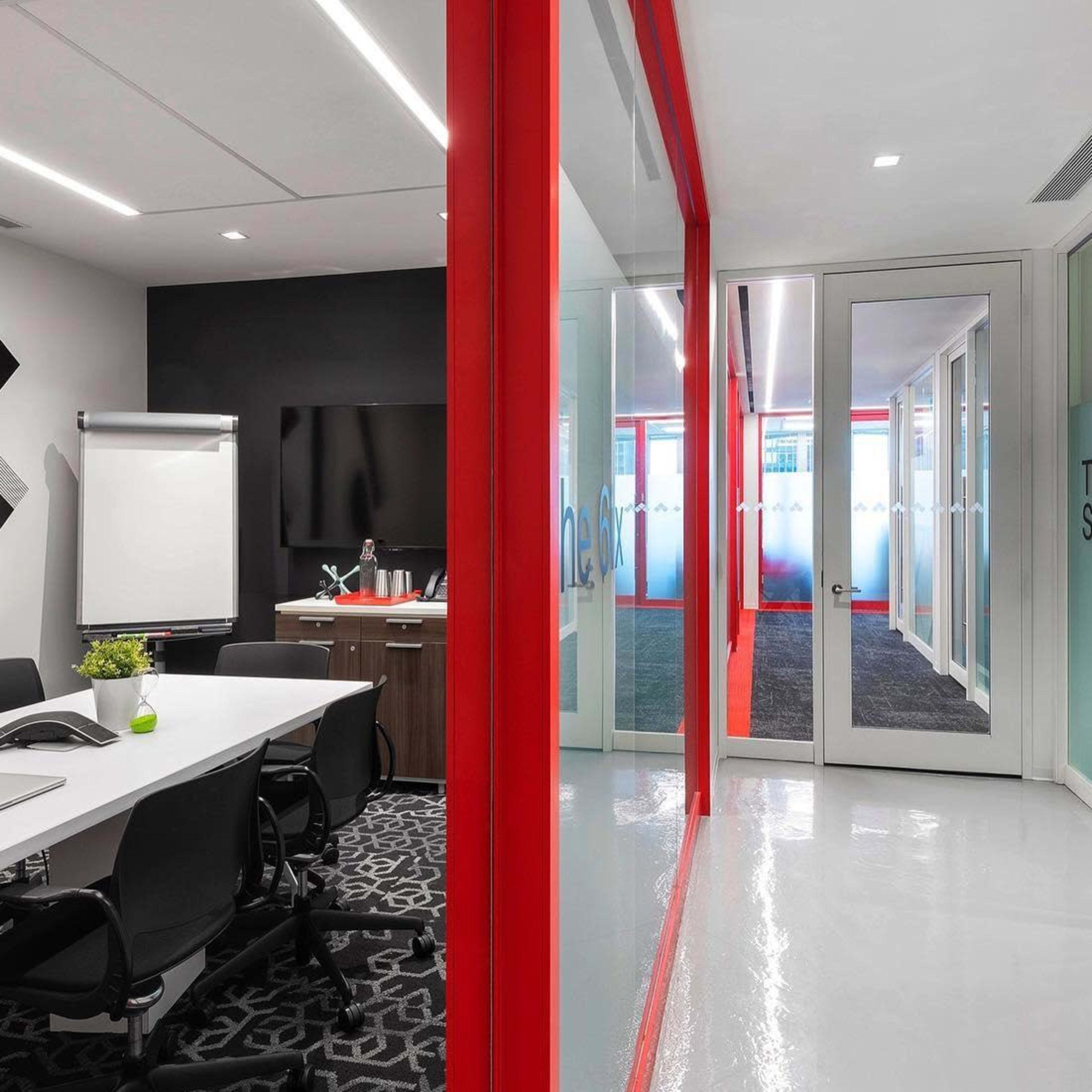A modern meeting room with a white table and black chairs, featuring a large screen and a glass wall leading to a hallway with red accents.