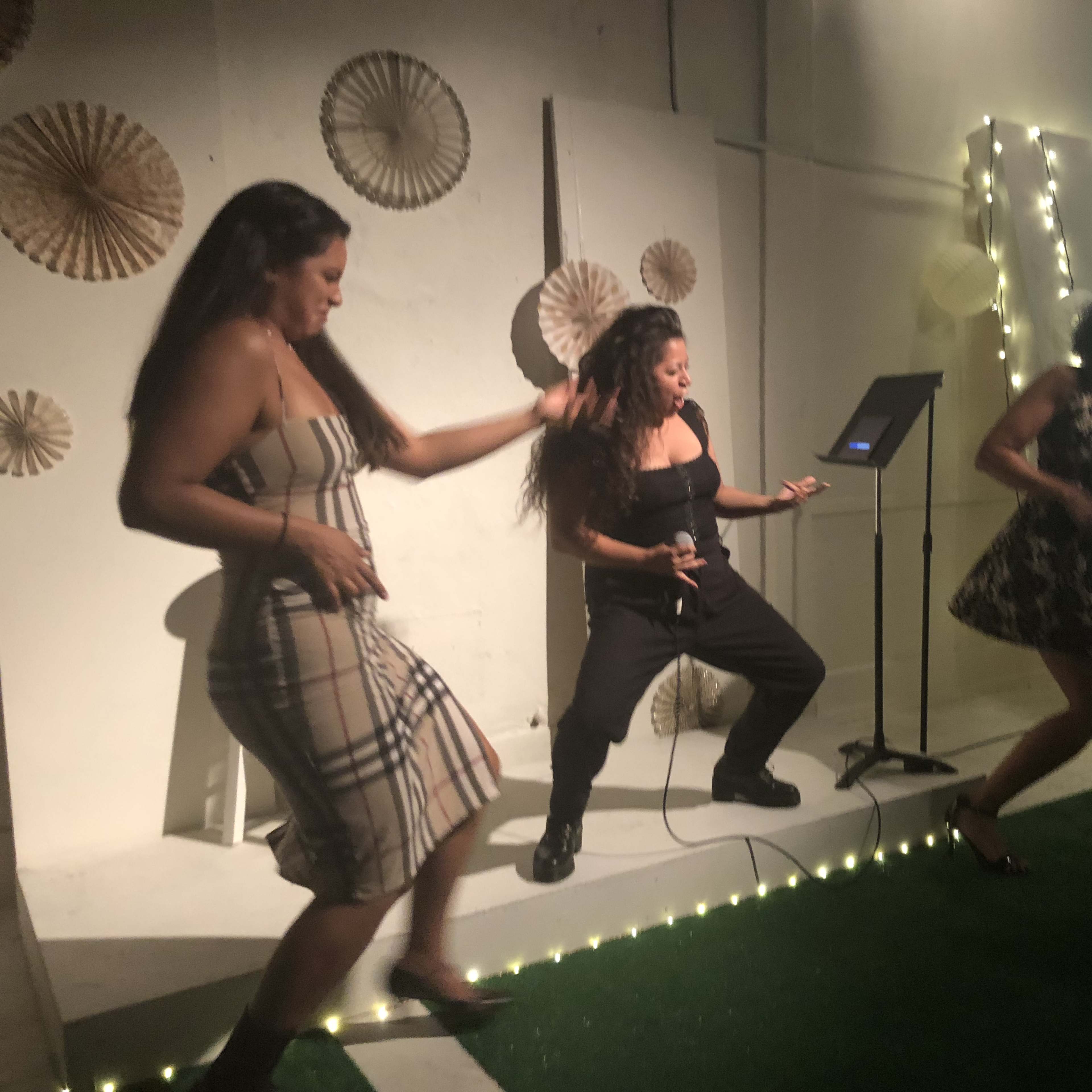 Three women perform a dance routine on stage with a microphone and a backdrop featuring a projected image.