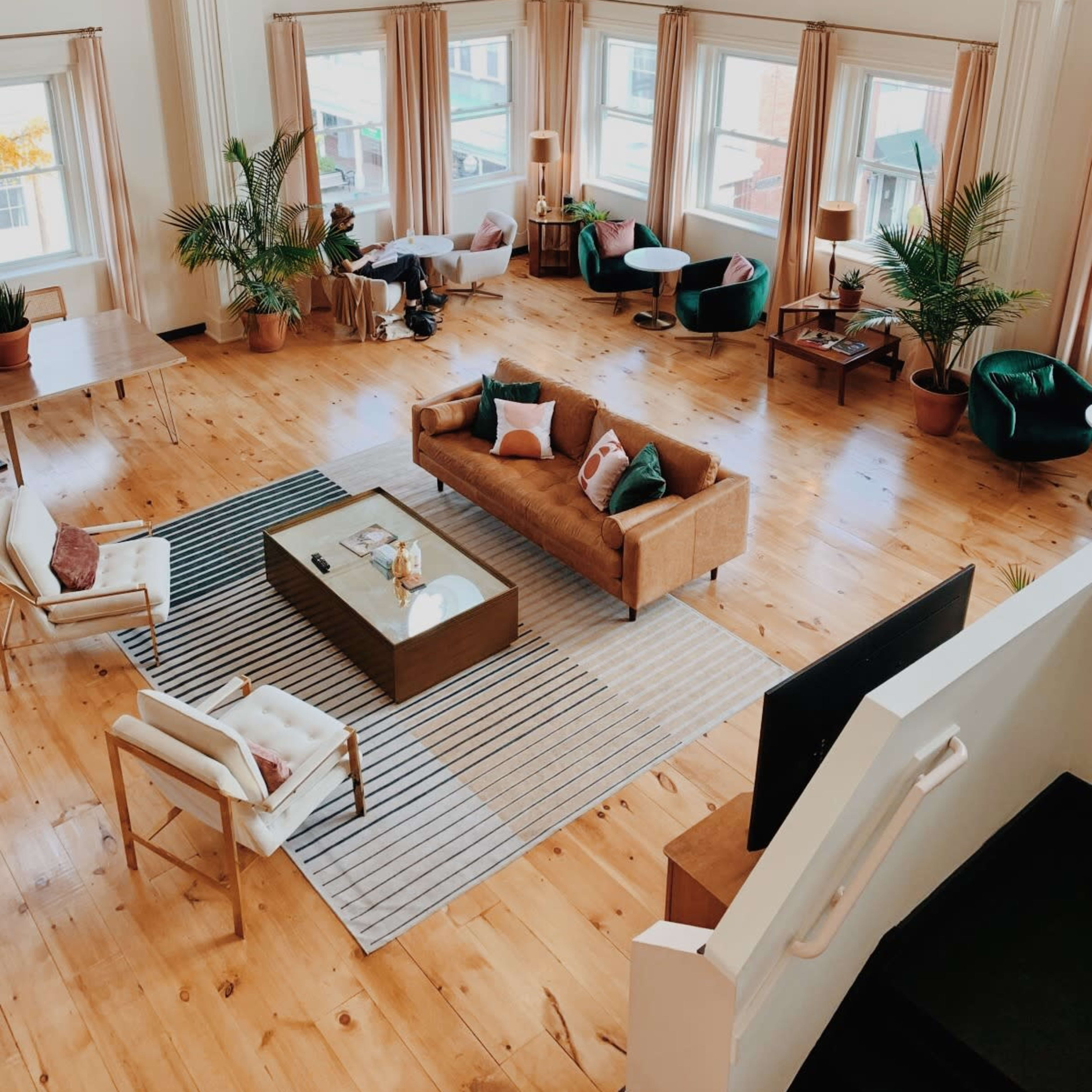 A spacious, elegantly designed living area with large windows, wooden flooring, and a combination of seating arrangements surrounded by plants.