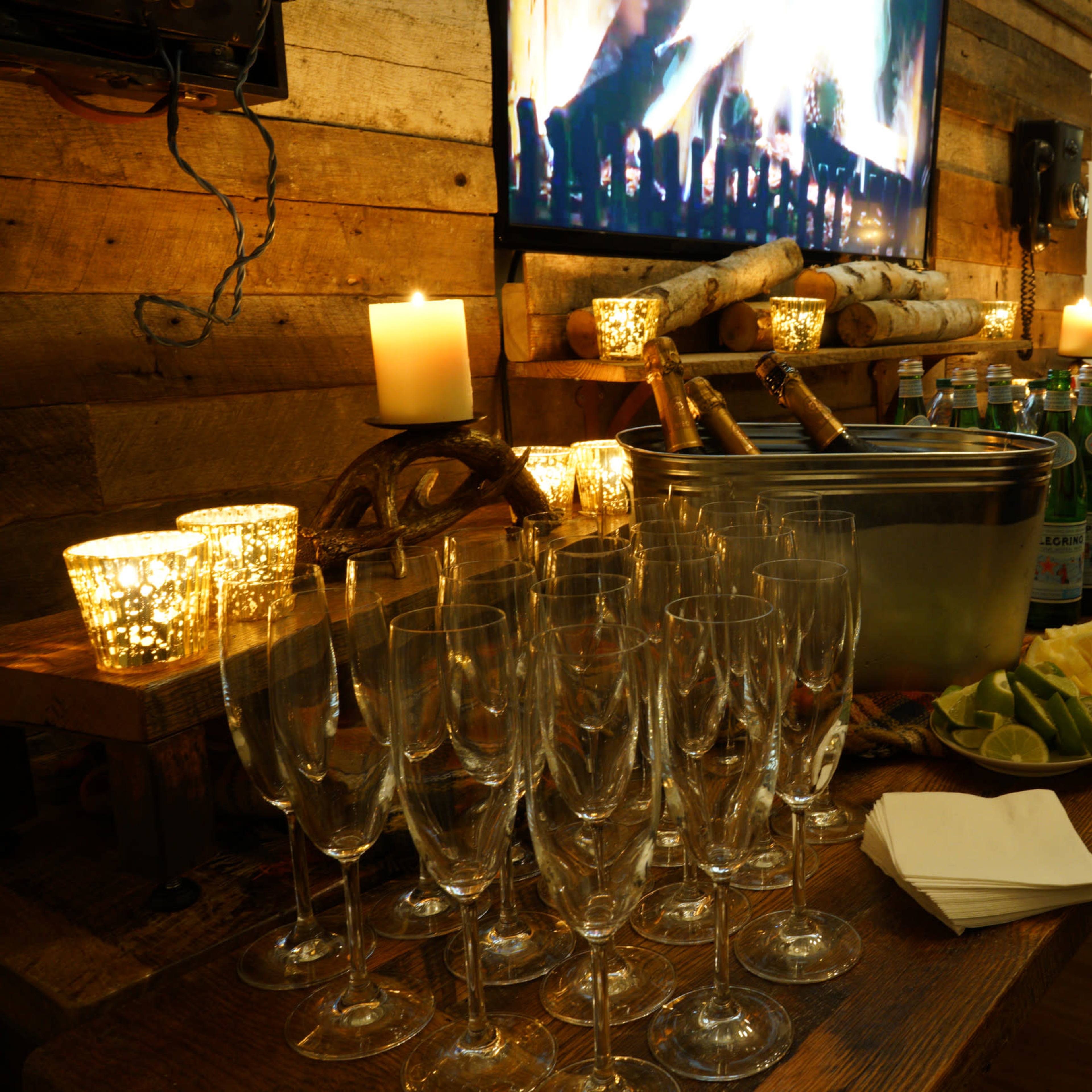 A wooden table is set with champagne flutes, candles, a large ice bucket with bottles, and refreshments, alongside a television displaying a festive scene.