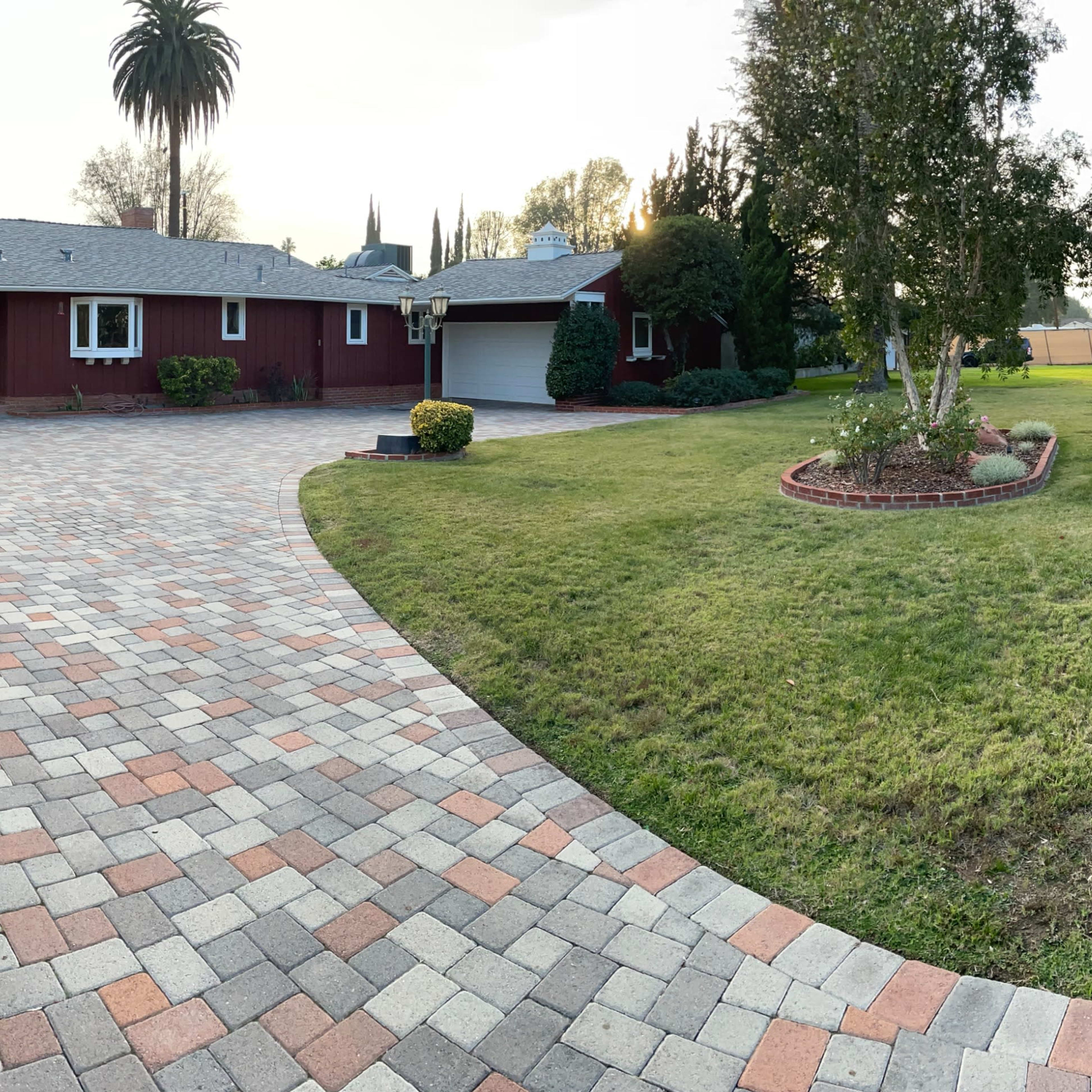 A residential front yard with a paved driveway leading to a house, surrounded by landscaped grass and bushes.
